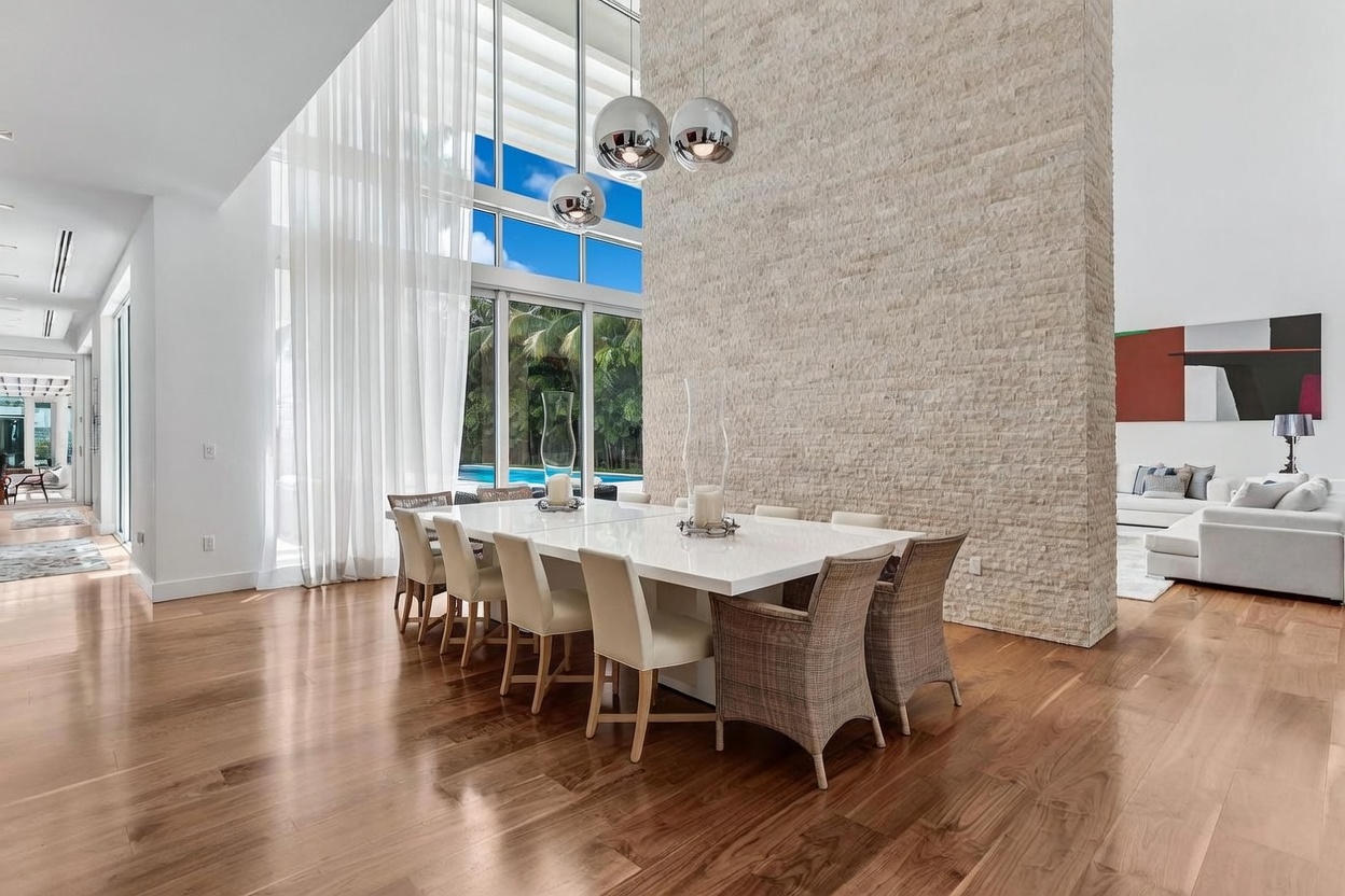 This bright and airy dining room features a large, glossy white table surrounded by modern chairs with woven backs. A prominent, textured stone wall serves as a central architectural feature, while tall windows with sheer curtains allow for abundant natural light and views of the outdoor pool area. The space is accented by a cluster of spherical pendant lights and polished wood flooring, creating a sophisticated and open atmosphere.