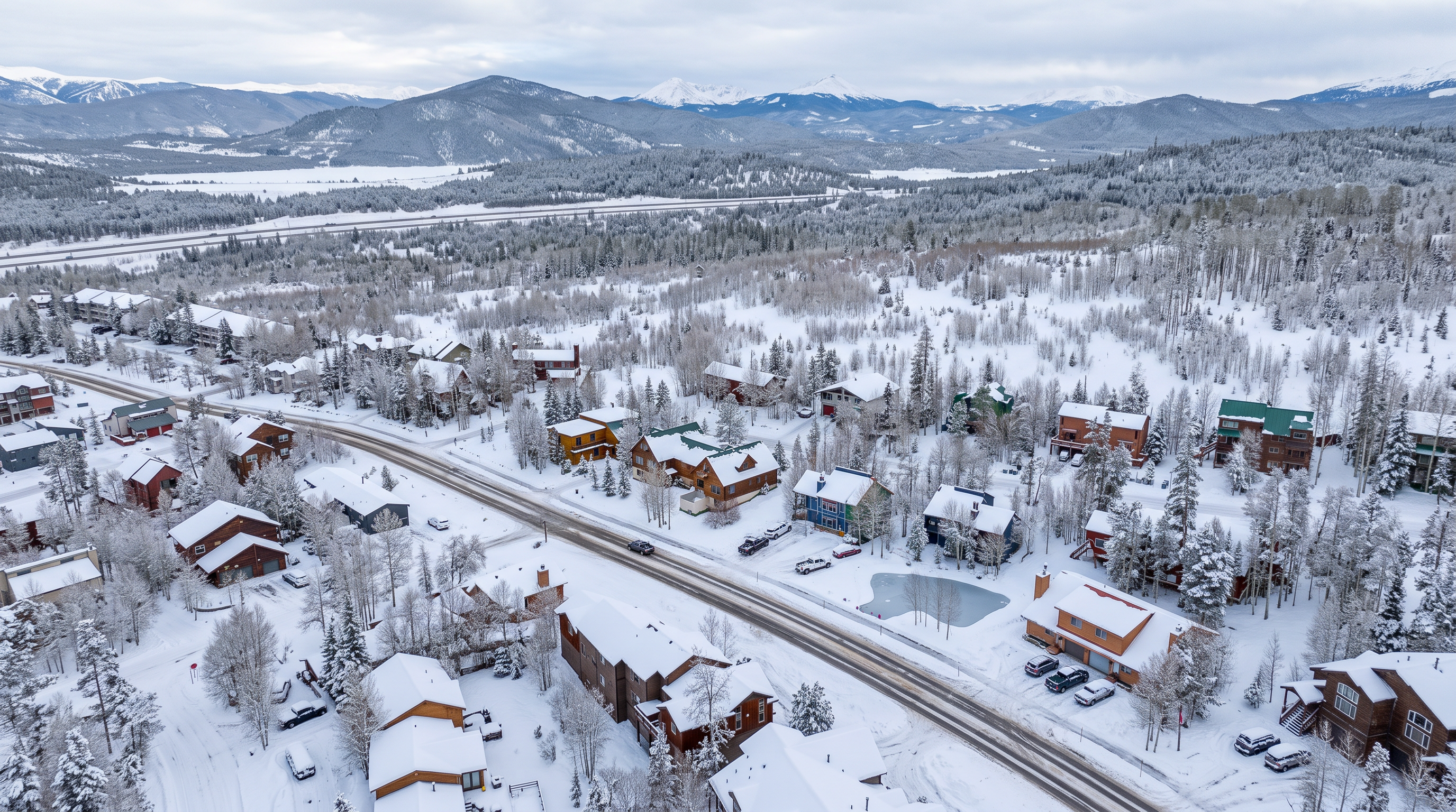 This high-altitude aerial shot captures a charming mountain residential neighborhood blanketed in fresh snow. The area features a variety of multi-level cabin-style homes surrounded by sparsely wooded snowy landscapes and distant, majestic snow-capped peaks. The scene offers a serene and picturesque view ideal for those looking for a tranquil retreat in a snowy, alpine environment.