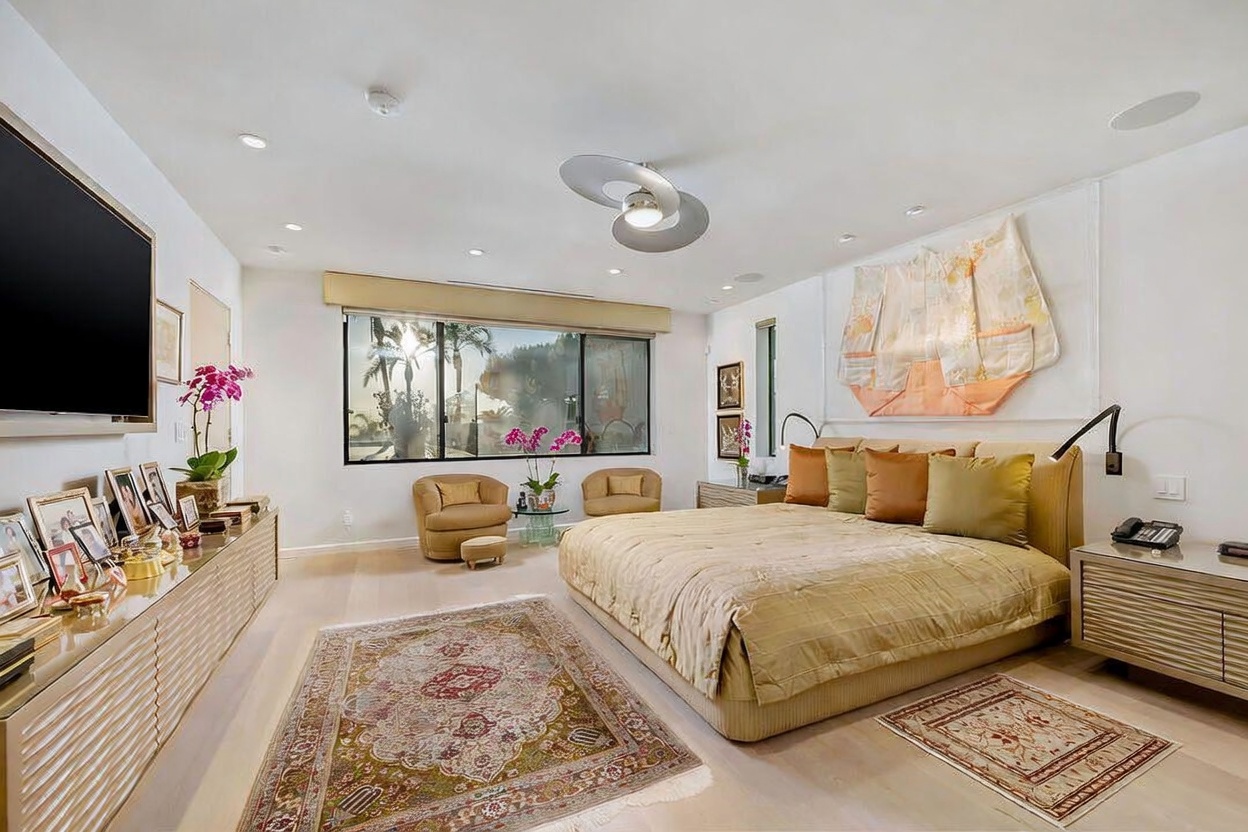 This spacious primary bedroom features a large bed with neutral-toned bedding and a decorative textile wall hanging above the headboard. The room is accented by a patterned area rug, two comfortable armchairs near the window, and a long, light-toned storage console beneath a wall-mounted television. Large windows allow natural light to fill the space, creating an airy and serene atmosphere.