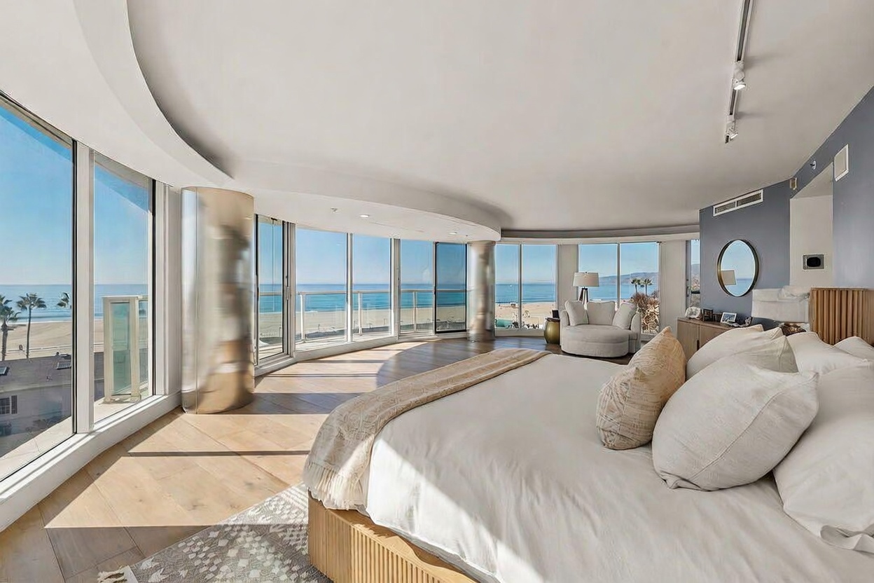 This primary bedroom features a curved wall of floor-to-ceiling windows that provide expansive, unobstructed views of the ocean and beach. The room is anchored by a large bed with neutral bedding and light wood flooring, creating a serene and airy atmosphere. A cozy seating area with a round chair is positioned near the glass, emphasizing the bright, coastal aesthetic of this luxury space.