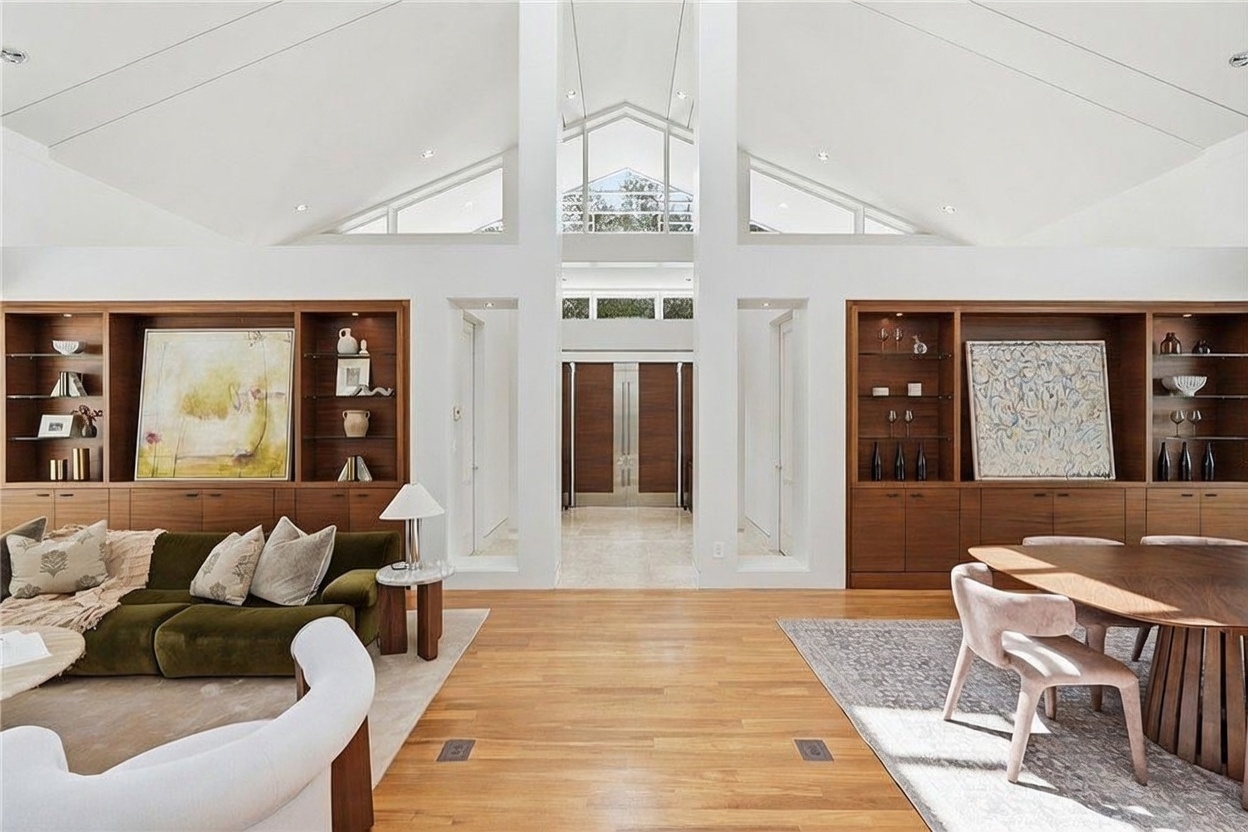 This expansive, open-concept living and dining area features a dramatic vaulted ceiling with large triangular windows that flood the space with natural light. The room is anchored by symmetrical, built-in wood shelving units on either side of a central entryway, creating a sophisticated and balanced aesthetic. A plush green sofa and a modern dining set are arranged on light wood flooring, emphasizing the airy, high-end architectural design of the home.