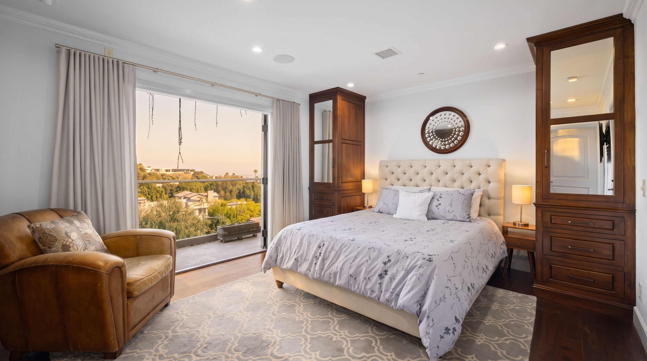 This primary bedroom features a tufted upholstered headboard centered between two tall, dark wood storage cabinets with integrated mirrors. A leather armchair sits near a large glass door that opens to a balcony, providing a scenic view of the surrounding landscape. The room is finished with dark wood flooring, a patterned area rug, and recessed lighting, creating a sophisticated and tranquil atmosphere.