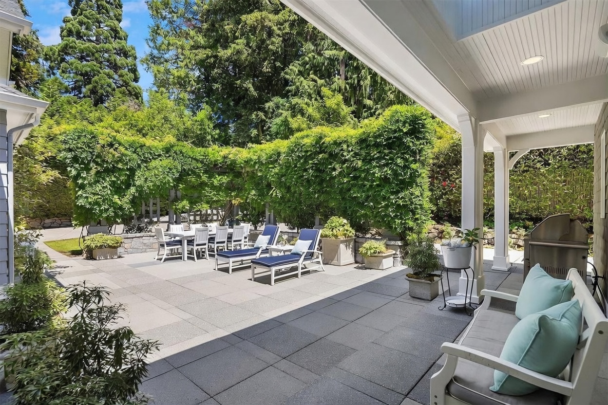 This spacious stone-paved patio offers an inviting outdoor living area, complete with a dining set and lounge chairs arranged under a lush, green-covered pergola. The space is partially covered by a roof overhang, providing a shaded seating area with a bench and a stainless steel grill nearby. The scene is framed by mature trees and dense greenery, creating a private and serene atmosphere perfect for entertaining.
