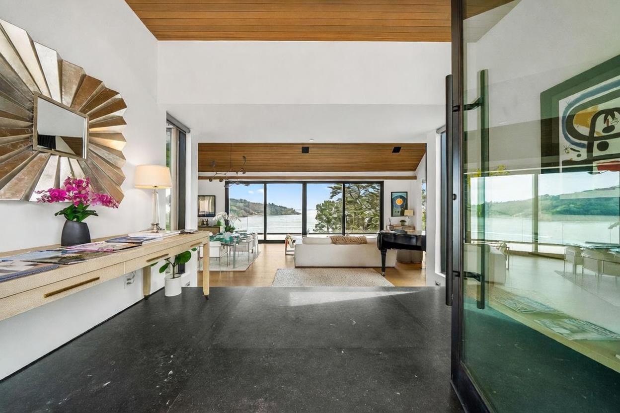This entryway offers a dramatic transition into a bright, open-concept living space with expansive views of the water through large glass walls. The foreground features a dark stone floor and a long, light-toned console table topped with a decorative mirror and vibrant flowers. The interior design emphasizes a seamless connection between the home's warm wood-paneled ceilings and the natural scenery outside.