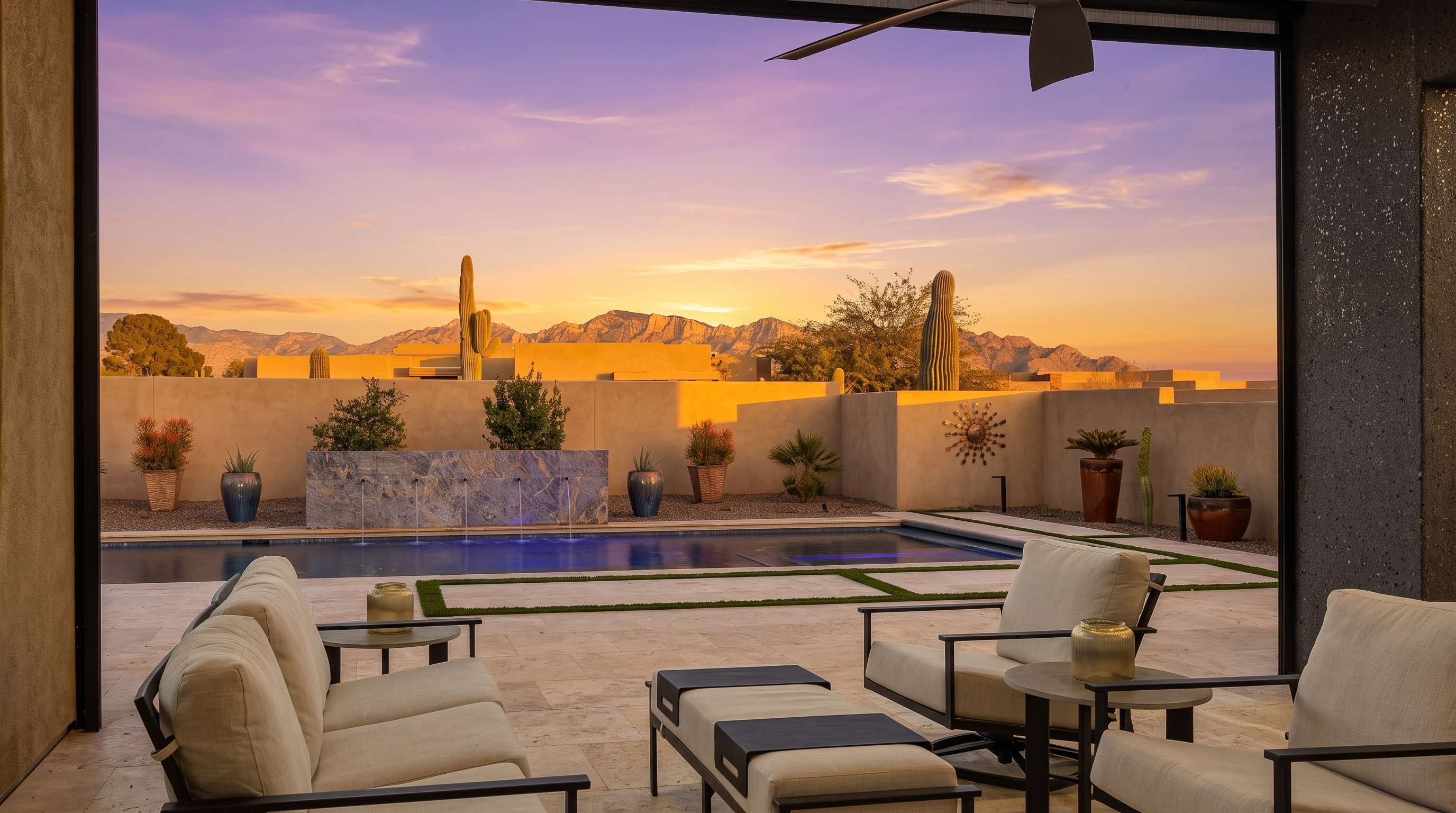 This covered patio offers a seamless transition to an outdoor oasis, featuring a modern seating area with light-toned cushions and dark metal frames. Beyond the patio, a sleek swimming pool with a blue-lit water feature serves as the focal point, set against a backdrop of desert landscaping and distant mountain ranges. The perspective is framed by the patio's overhead structure, creating a cinematic, open-air feel that highlights the property's connection to the surrounding landscape.