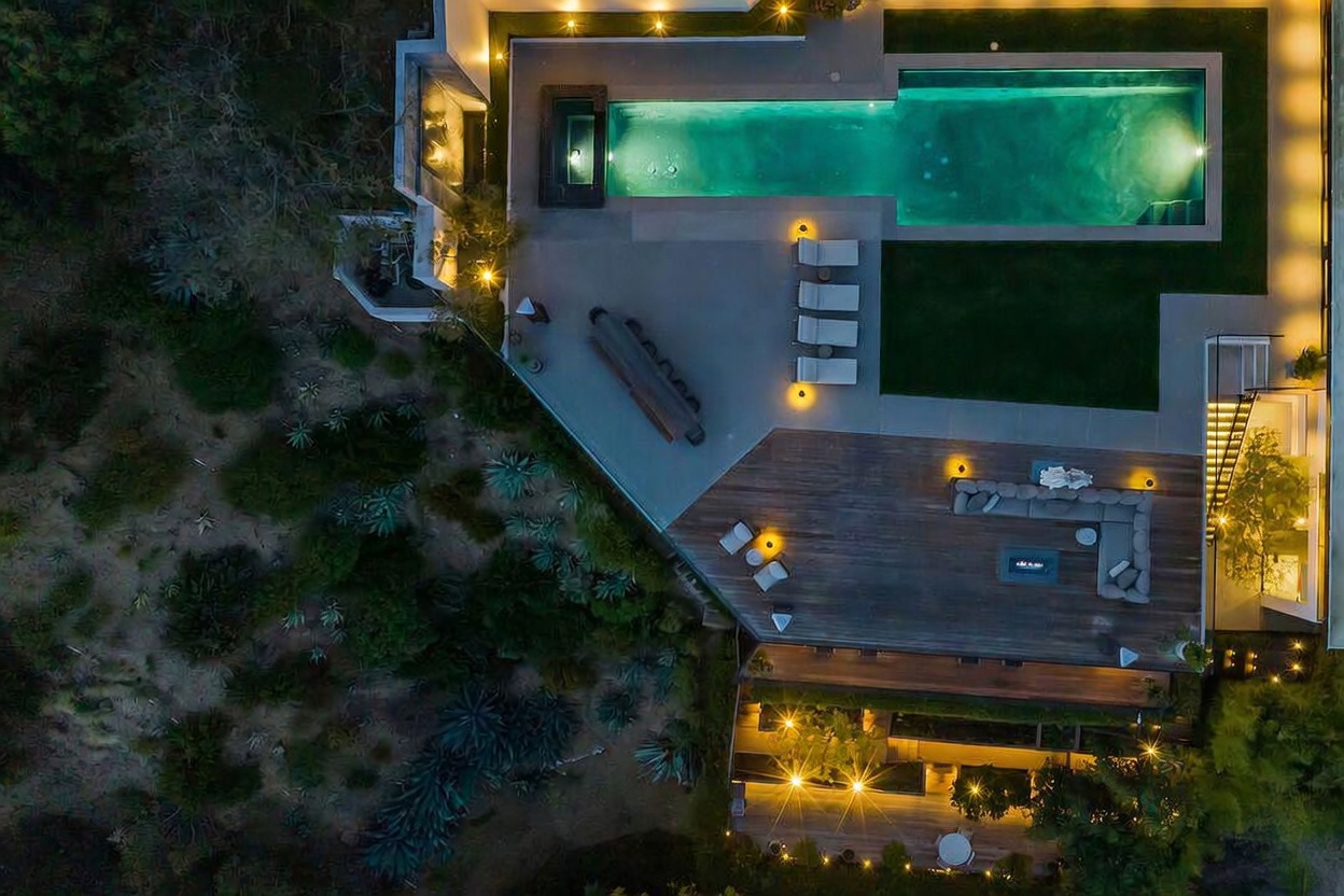 This high-angle aerial view captures a luxurious modern outdoor living space at dusk, featuring a long, illuminated rectangular swimming pool and an adjacent spa. A spacious wooden deck is furnished with a large sectional sofa and a fire feature, while a separate paved patio area includes a row of lounge chairs. The property is seamlessly integrated into a natural, landscaped hillside, creating a sophisticated and serene atmosphere perfect for evening entertaining.