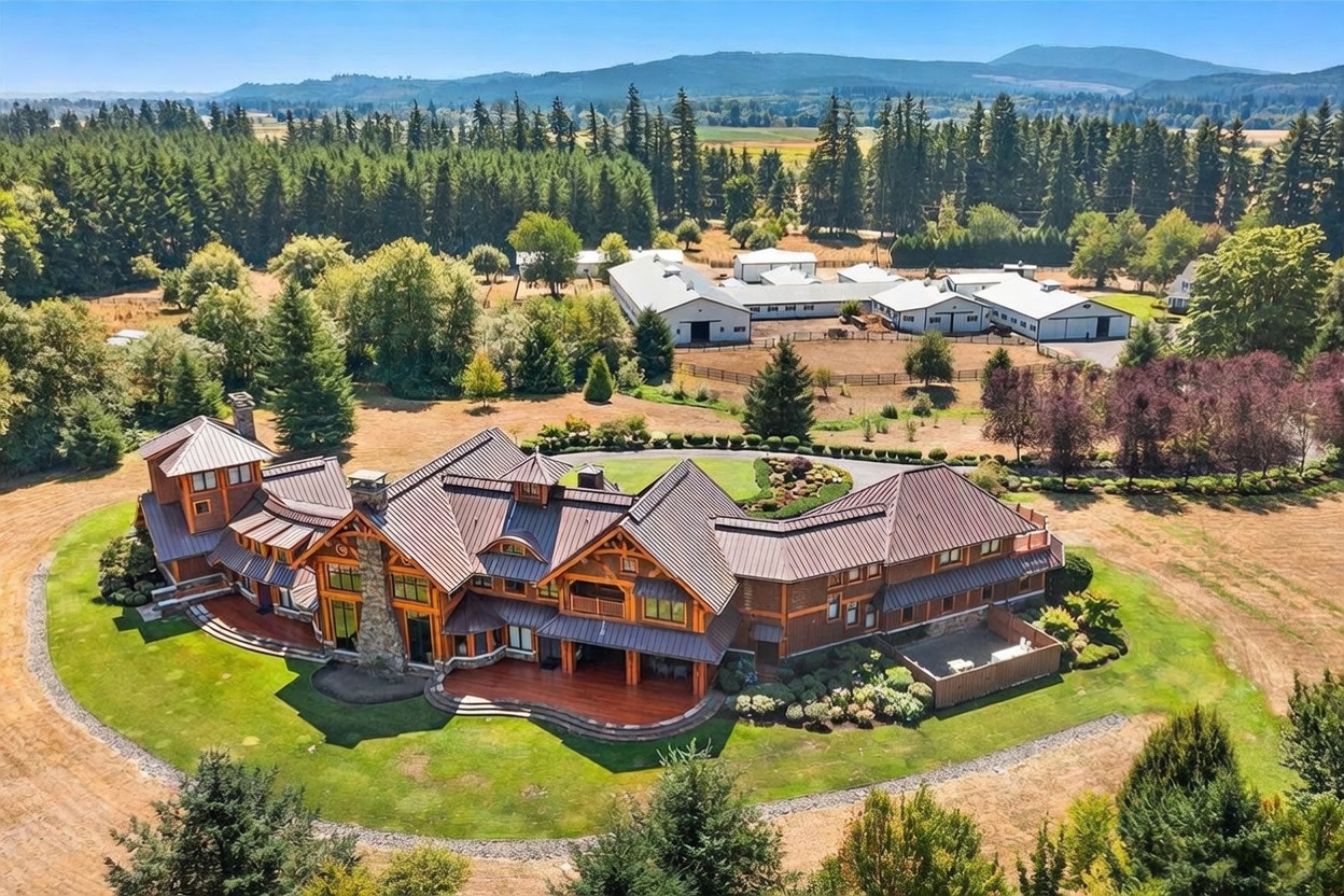 This high-angle aerial view captures a sprawling, multi-level estate designed in a rustic, lodge-inspired architectural style with dark metal roofing and wood-paneled siding. The property features a large, curved wooden deck overlooking a manicured lawn, with a stone-clad chimney breast rising prominently along the rear facade. In the background, a complex of large outbuildings and barns sits amidst a vast landscape of trees and open fields, emphasizing the expansive nature of the rural estate.