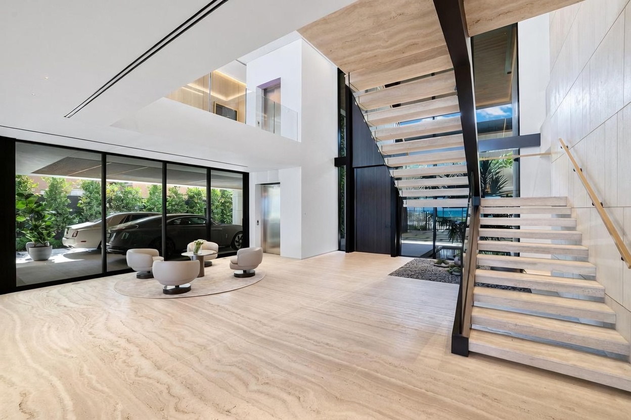 This expansive, double-height foyer features a dramatic floating staircase with light wood treads and a glass railing, serving as the architectural centerpiece of the space. To the left, a large glass wall provides a clear view into an attached garage housing luxury vehicles, while a minimalist seating area sits on a circular rug in the foreground. The room is finished with polished stone flooring and clean white walls, creating a bright, gallery-like atmosphere that emphasizes modern luxury and open-concept design.