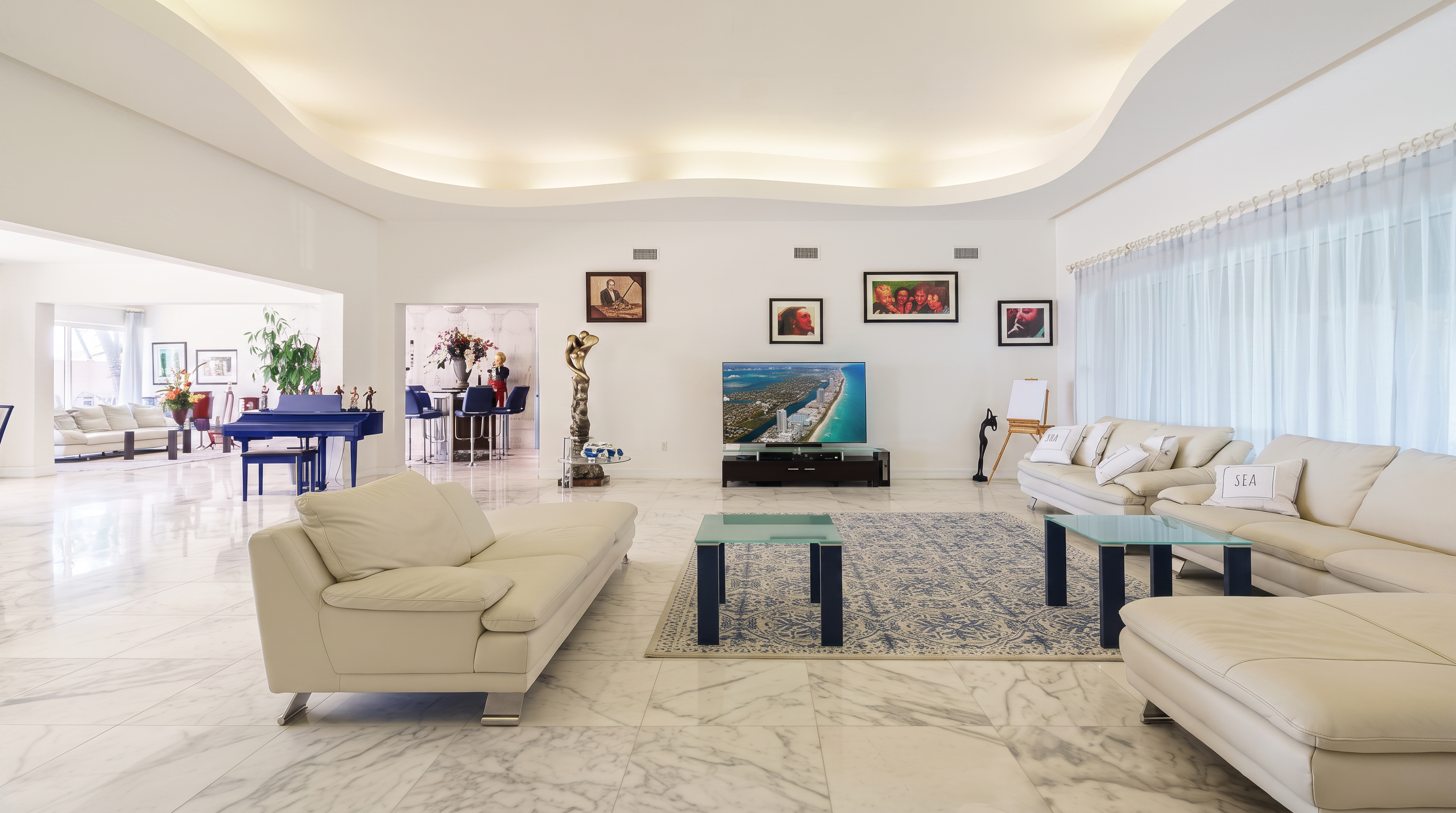 This spacious, open-concept living room features bright white walls, recessed cove lighting, and polished stone flooring. The room is furnished with large, light-colored sectional sofas arranged around a patterned area rug, with a central television console and several framed artworks displayed on the walls. A blue grand piano is visible in the background, adding a touch of elegance to the airy, well-lit living space.