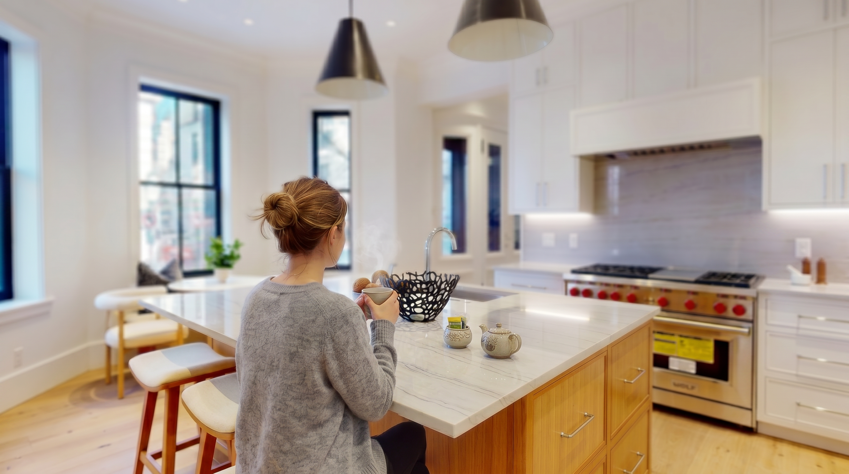 This bright, modern kitchen features a large central island with a light-toned stone countertop and integrated seating, complemented by warm-toned wood cabinetry. The space is anchored by white upper cabinets, a professional-grade stainless steel range, and two prominent pendant lights hanging above the island. Large windows with dark frames provide ample natural light and a view of the street, creating an airy and inviting atmosphere.