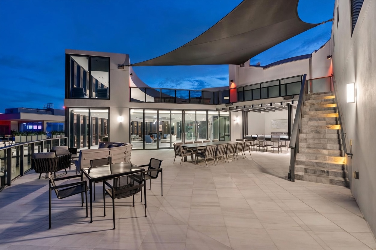 This expansive, modern rooftop terrace features a large dining area with a long table and multiple chairs, alongside a comfortable lounge seating arrangement. The space is partially covered by a large, sleek shade sail and includes a built-in outdoor bar area, with a stone staircase leading to an upper level. Large glass doors provide a seamless transition to the interior, creating a sophisticated and airy atmosphere perfect for evening entertaining.
