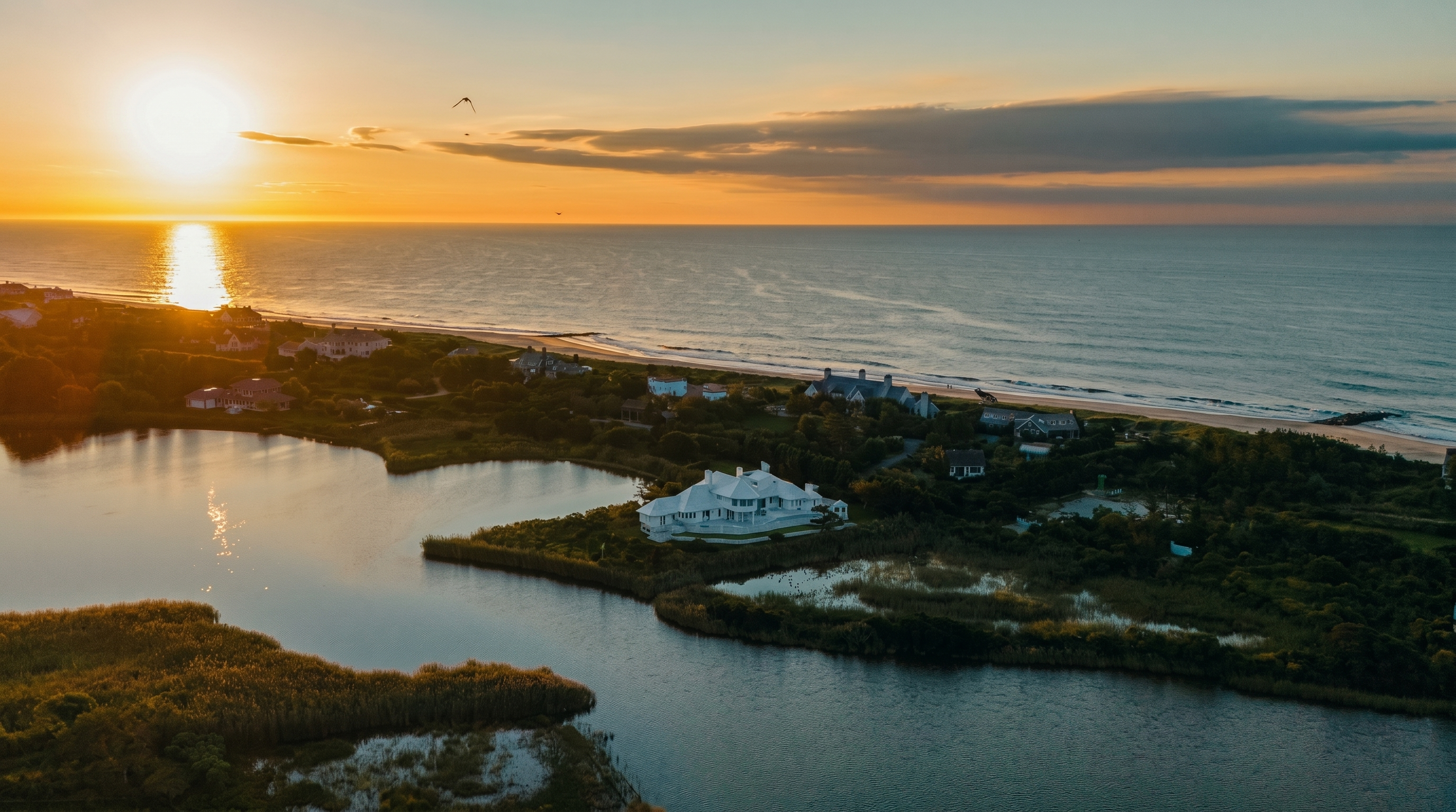 This stunning aerial perspective captures a luxurious white estate situated on a peninsula between a tranquil pond and the vast ocean. The property features expansive grounds, lush greenery, and a prime waterfront location that emphasizes its secluded and prestigious setting. The golden hour lighting casts a warm, cinematic glow over the entire landscape, highlighting the serene coastal atmosphere.