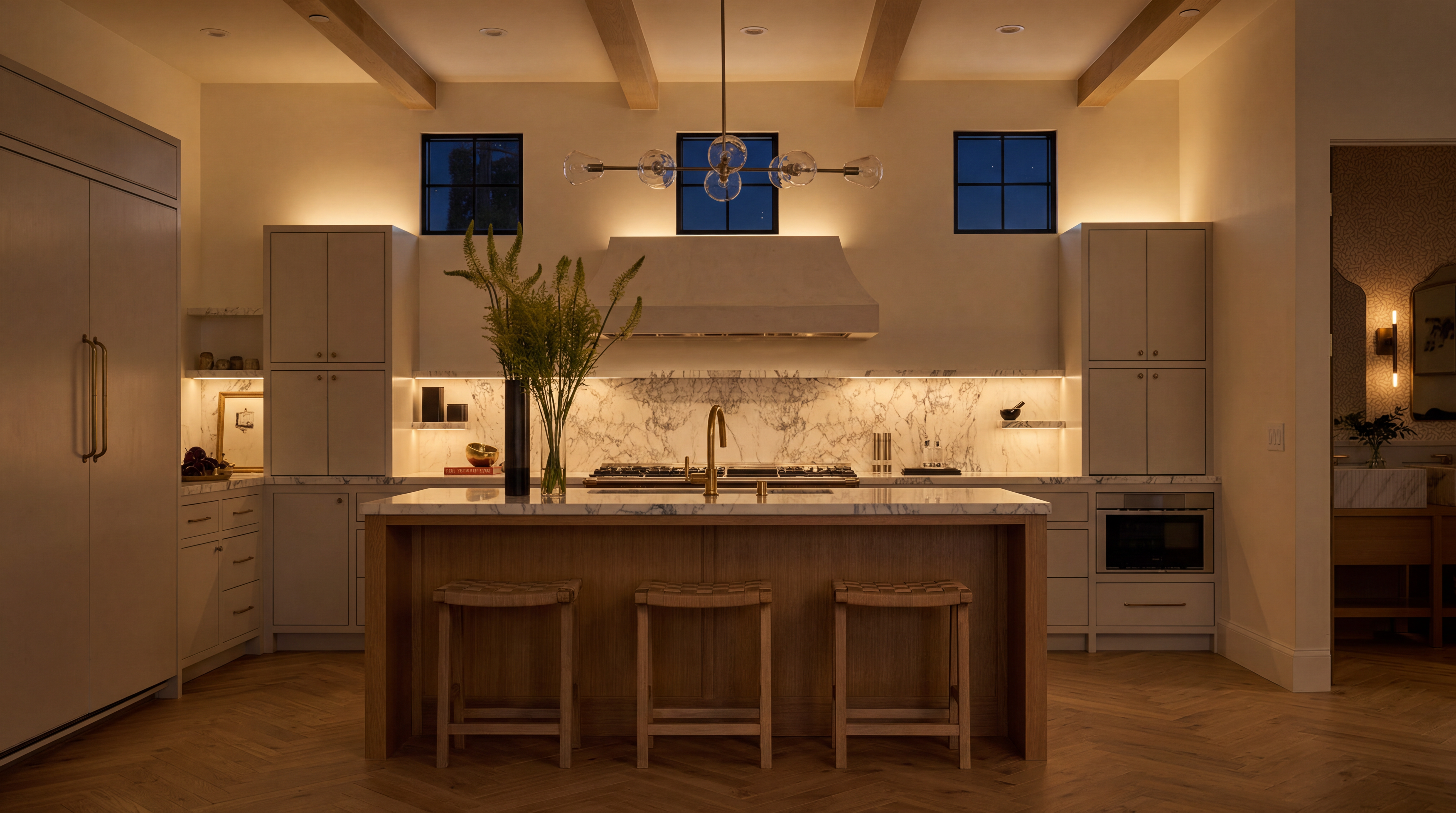 This sophisticated kitchen features high-end finishes including marble backsplash and countertops, warm wood chevron flooring, and custom cabinetry with integrated appliances. A large central island with woven bar stools provides ample workspace and seating under a modern chandelier, while bespoke recessed and under-cabinet lighting creates an inviting, upscale ambiance. The layout is balanced and elegant, showcasing a seamless blend of modern function and timeless design.