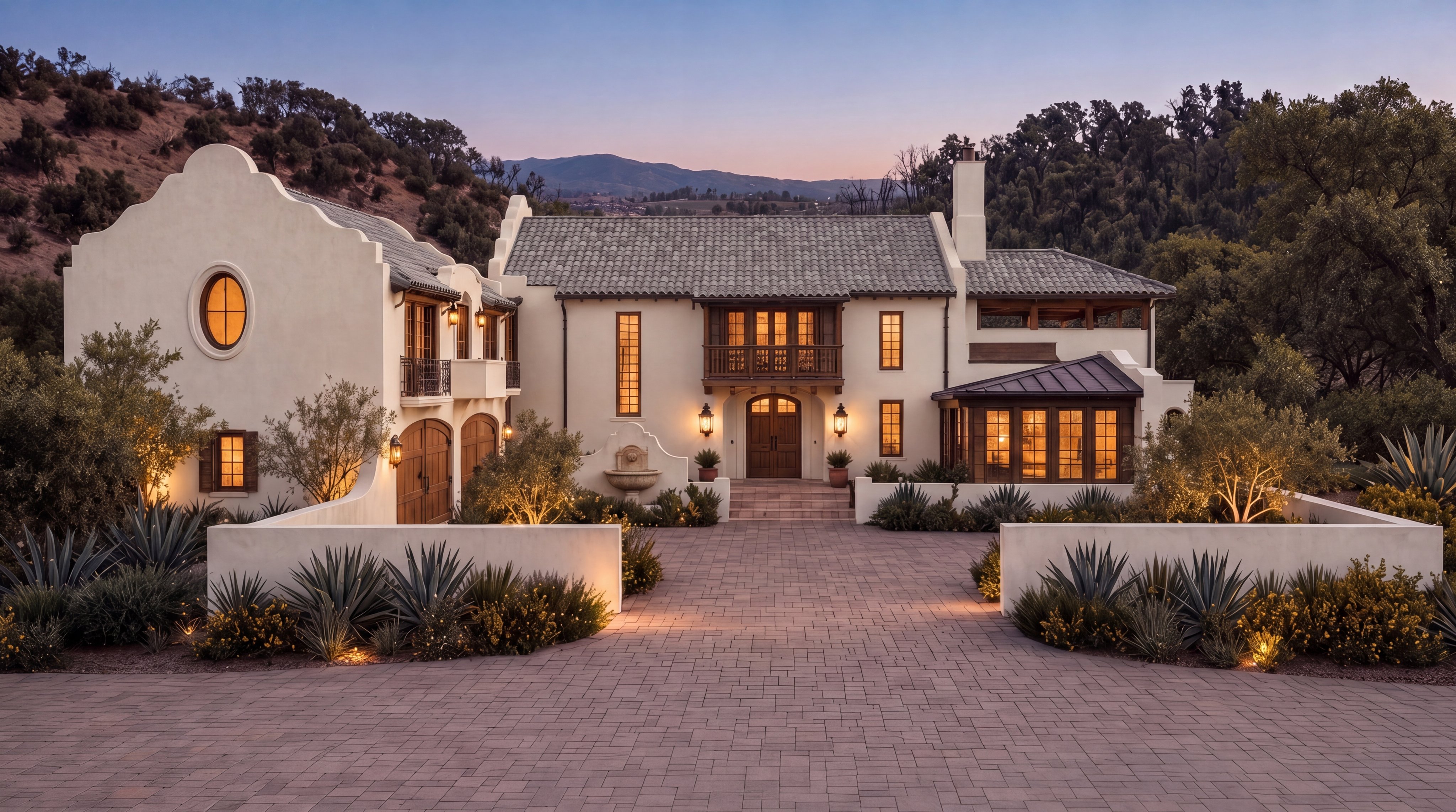 This stunning Mediterranean-style estate features a symmetrical stucco façade with a tile roof and charming arched architectural details. The property is highlighted by a beautifully paved grand driveway, warm ambient exterior lighting, and lush drought-tolerant landscaping including agave plants. Its elegant curb appeal is further enhanced by a rustic wooden front door, a balcony, and a scenic backdrop of rolling hills at dusk.