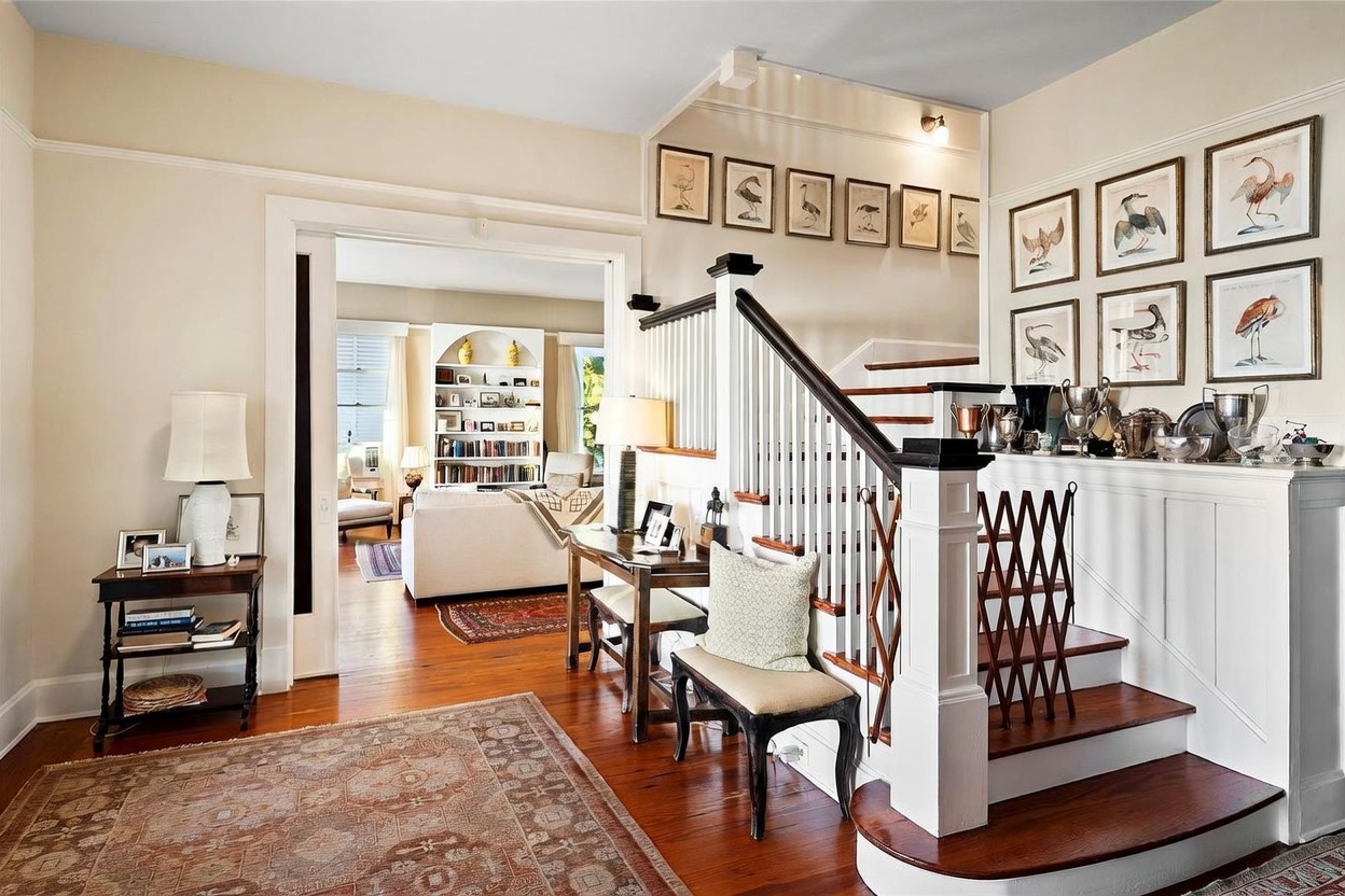 This elegant foyer features warm wood flooring and a classic staircase with a dark wood handrail and white balusters. A gallery wall of framed bird prints adds character to the space, while a small console table and bench provide functional seating. The perspective looks through a wide doorway into a bright, inviting living area, creating a sense of depth and flow throughout the home.