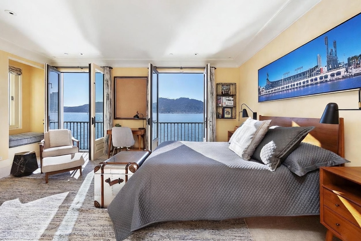 This primary bedroom features a serene, light-filled atmosphere with direct access to a private balcony overlooking the water through large glass doors. The room is furnished with a quilted gray bed, a vintage-style trunk at the foot of the bed, and a cozy seating area near the window. A large panoramic photograph of a stadium hangs above the headboard, adding a personal touch to the neutral, sun-drenched space.