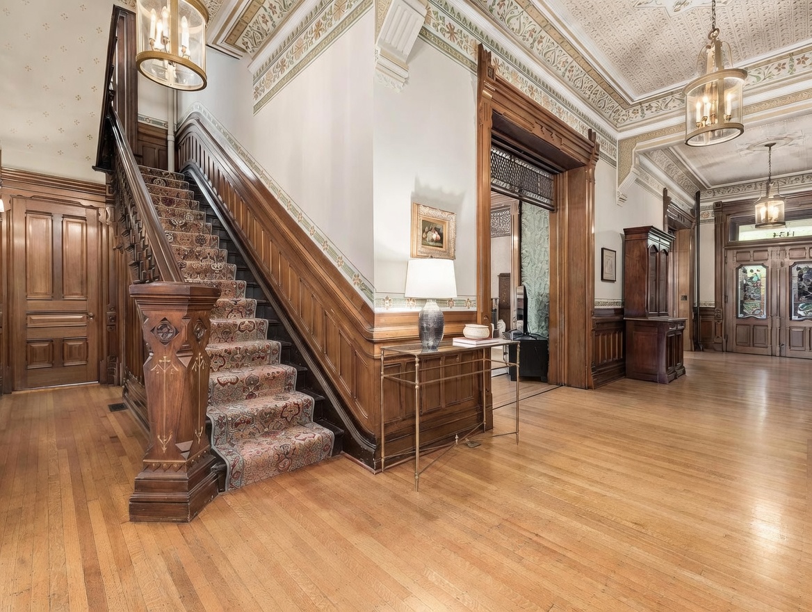 This grand foyer showcases historic architectural details, including an ornate wooden staircase with a patterned runner and intricate carved newel posts. The space features polished wood flooring, high ceilings with decorative molding, and warm ambient lighting from elegant hanging lanterns. A slim metal console table sits against the wall, leading the eye toward a wide, wood-trimmed doorway that opens into an adjacent room.