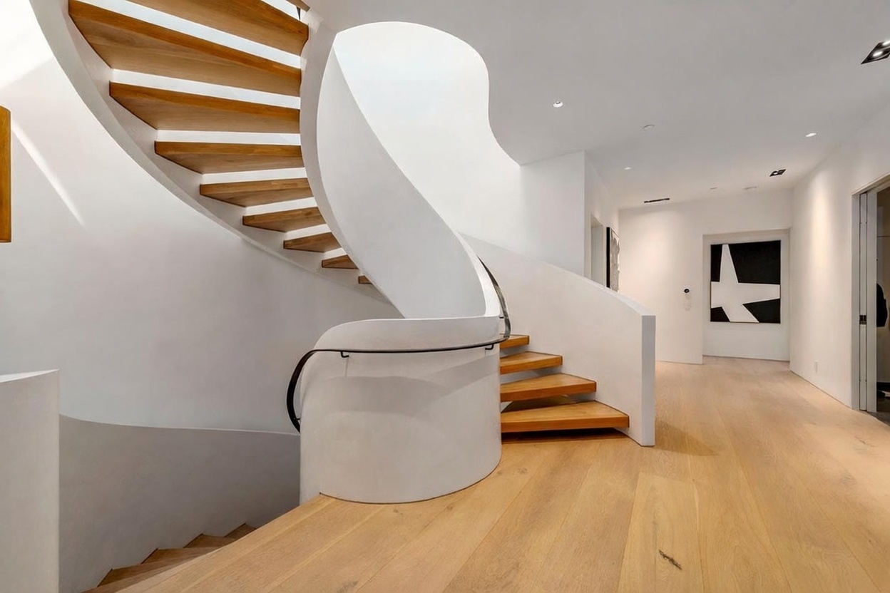 This image captures a striking, sculptural spiral staircase with light wood treads and a smooth, white curved wall. The minimalist design creates a bright and airy atmosphere, complemented by light wood flooring that extends into the adjacent hallway. A piece of abstract black-and-white art hangs on the far wall, adding a modern focal point to the clean, architectural space.