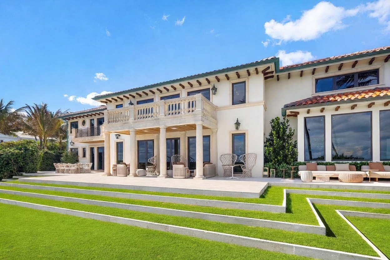 This expansive rear view of a Mediterranean-style estate showcases a multi-level stone patio furnished with elegant outdoor seating areas. The home features a prominent second-story balcony with stone balustrades, large glass doors, and a classic tile roof, all overlooking a tiered lawn with geometric concrete retaining walls. The bright, sunny atmosphere highlights the architectural grandeur and the seamless transition between the indoor living spaces and the manicured backyard.