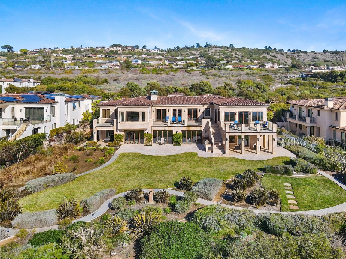 This aerial perspective captures the expansive rear elevation of a multi-story Mediterranean-style residence, showcasing a symmetrical design with a tiled roof and light-toned stucco walls. The home features multiple balconies and large glass doors that open onto a stone patio, which transitions into a manicured lawn and landscaped garden. The property is set against a backdrop of rolling hills, emphasizing its private and scenic location.