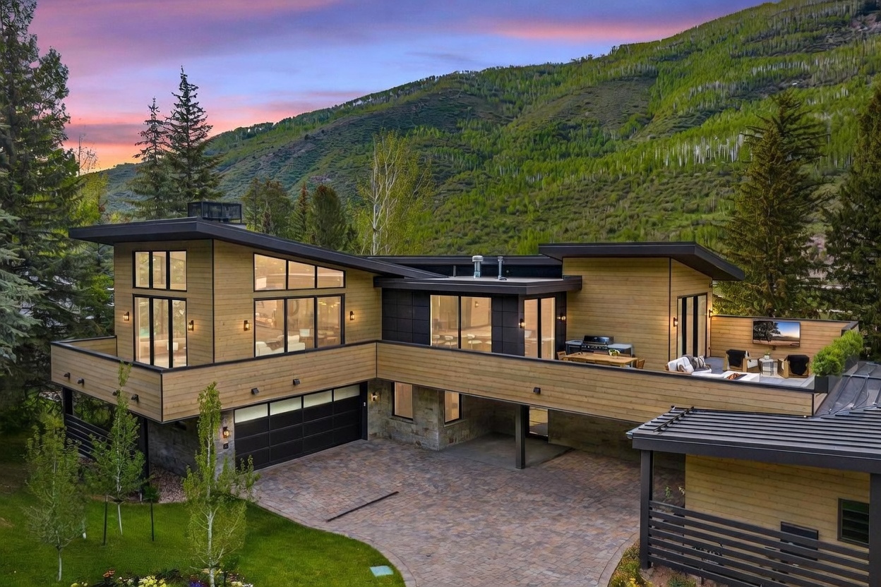 This modern mountain residence features a striking architectural design with a multi-level layout, horizontal wood siding, and dark metal accents. The home includes a spacious elevated deck with an outdoor kitchen and lounge area, overlooking a paved driveway and a lush, tree-lined landscape. The perspective is captured from an elevated angle, showcasing the home's integration into the hillside during a vibrant sunset.