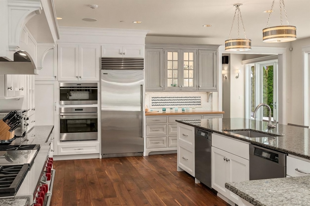 This bright, high-end kitchen features a blend of white and soft gray cabinetry paired with dark stone countertops and rich wood flooring. The space is equipped with professional-grade stainless steel appliances, including a large refrigerator, double wall ovens, and a dishwasher integrated into the central island. Two decorative pendant lights hang above the island, while a patterned tile backsplash adds a subtle design element to the prep area.