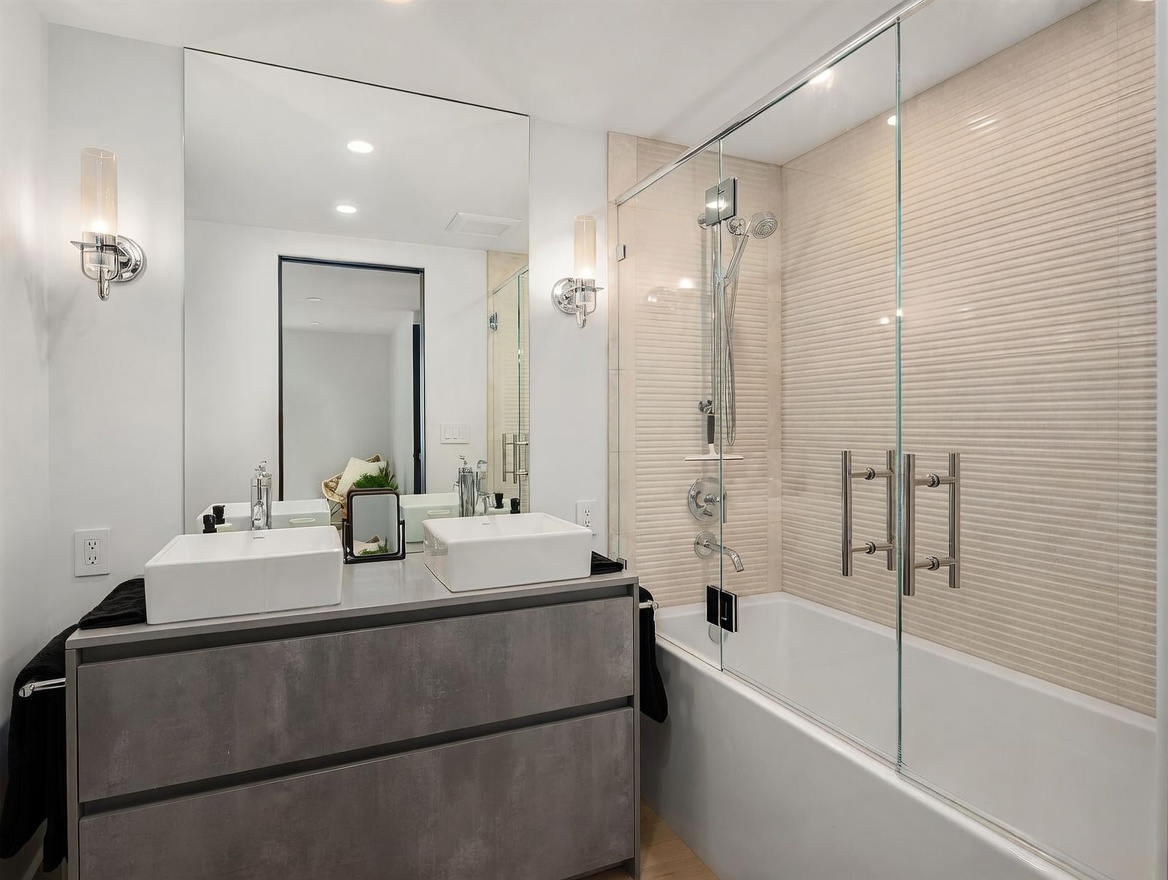 This modern bathroom features a double vanity with two square vessel sinks set atop a dark, textured cabinet. A large, frameless mirror spans the wall above the vanity, flanked by two sleek wall-mounted light fixtures. To the right, a glass-enclosed shower and tub combination is finished with horizontal textured wall tiles, creating a clean and contemporary aesthetic.