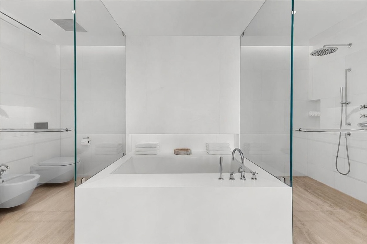 This modern, minimalist bathroom features a central soaking tub as the focal point, flanked by glass-enclosed shower and toilet areas. The space is finished with clean white surfaces and light wood-toned flooring, creating a bright and airy atmosphere. A wall-mounted faucet serves the tub, while the shower area includes a rainfall showerhead and integrated shelving.
