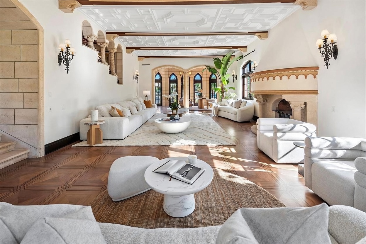 This expansive living room features a sophisticated, neutral palette with intricate coffered ceilings and rich, patterned wood flooring. The space is anchored by a large, curved stone fireplace and multiple seating areas, including plush white sofas and a central marble-topped coffee table. Tall, arched doorways lead to an adjacent sunlit area, creating an open and airy atmosphere with a grand, Mediterranean-inspired aesthetic.