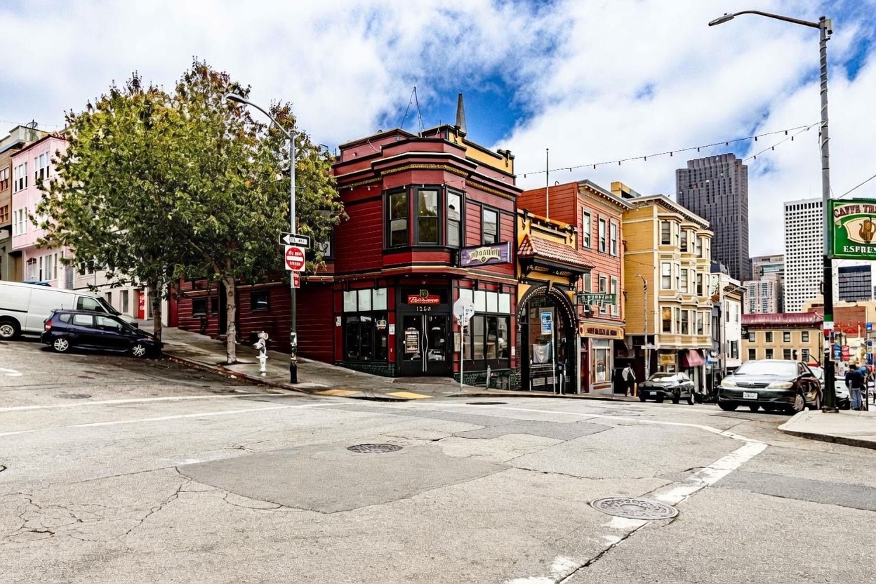 This street-level perspective captures a vibrant, multi-story commercial building on a corner lot in a dense urban setting. The structure features a distinct red-painted facade with a prominent bay window, adjacent to a row of colorful, classic architectural townhomes. The scene is set on a sloped street with a clear view of the surrounding neighborhood and distant city skyscrapers under a bright, cloudy sky.