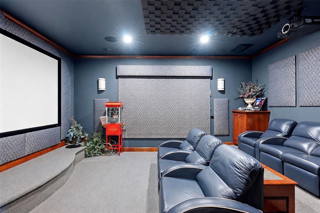 This dedicated home theater room features a large projection screen, tiered seating with blue leather recliners, and acoustic wall panels for an immersive viewing experience. A vintage-style popcorn machine adds a playful touch to the space, while the dark color palette and recessed lighting create a cozy, cinematic atmosphere. The room is designed for entertainment, complete with a small wooden cabinet for media storage and a projector mounted on the ceiling.