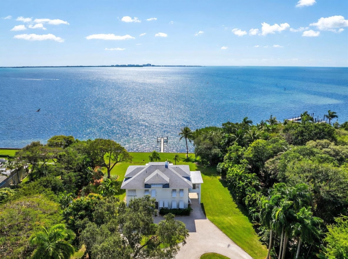 This high-angle aerial view captures a stately, multi-story residence situated on a lush, expansive waterfront lot. The property features a private dock extending into the calm, blue bay, surrounded by dense tropical landscaping and manicured green lawns. The home itself showcases a classic architectural style with a light-colored exterior, a multi-pitched roof, and a circular driveway leading to the front entrance.