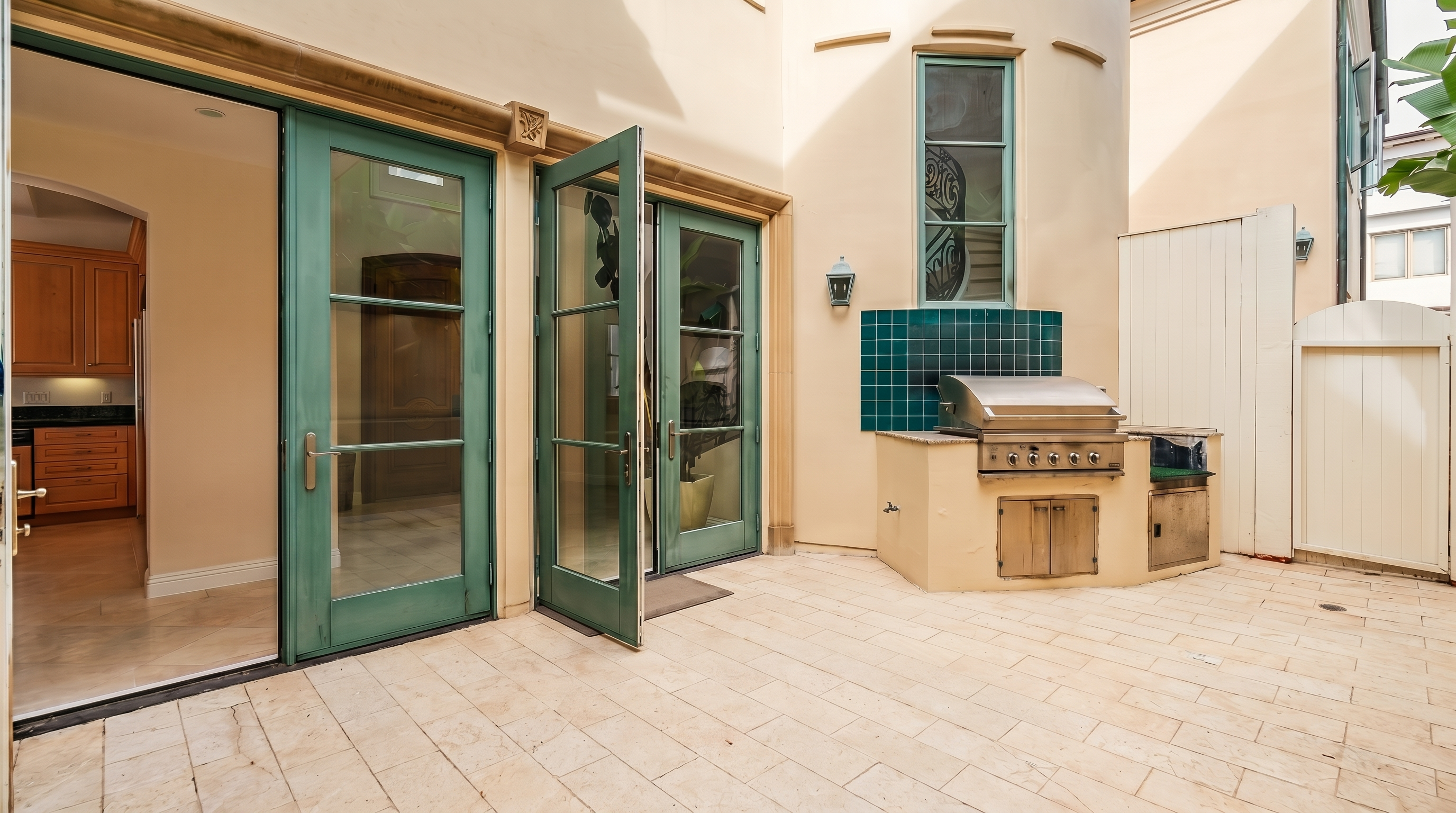 This outdoor patio space features light-colored stone pavers and a built-in outdoor kitchen area with a stainless steel grill. The area is accessed through double glass doors that open into an interior space, creating a seamless transition between indoor and outdoor living. The architectural style includes a curved wall section and a tall, narrow window, providing a bright and airy atmosphere.