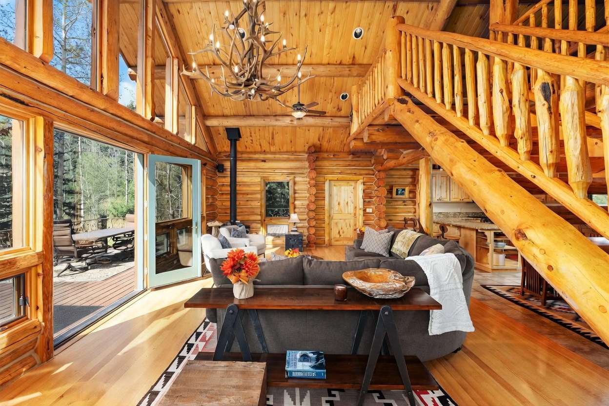 This rustic living room features exposed log walls, a vaulted wood-paneled ceiling, and a prominent antler chandelier. A large sectional sofa faces a wood-burning stove, while a staircase with log railings leads to an upper level. Large windows and a glass door provide abundant natural light and a view of the surrounding forest.