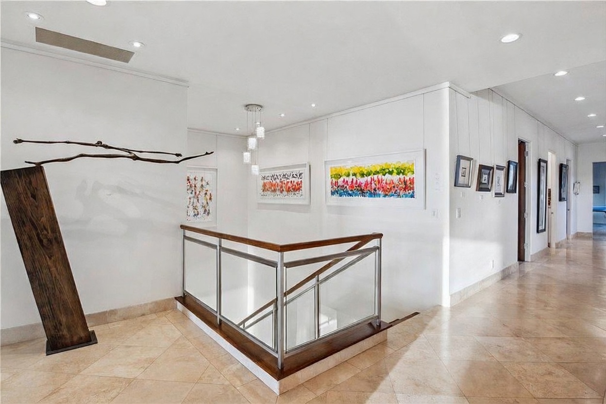 This bright, gallery-style hallway features a staircase with a modern glass and wood railing, leading to a lower level. The space is adorned with vibrant, colorful artwork on the white walls and illuminated by recessed ceiling lights and a contemporary multi-pendant fixture. The floor is finished with large, light-toned stone tiles, creating a clean and sophisticated atmosphere.