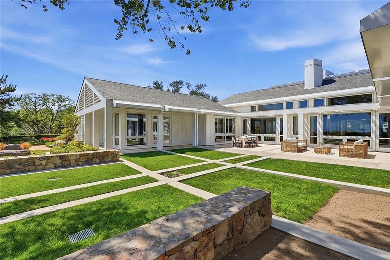 This rear view of a modern, single-story residence showcases a sprawling layout with extensive glass walls and a clean, white exterior. The home opens onto a manicured lawn divided by stone pathways, leading to a paved patio area furnished with outdoor seating. The architectural style emphasizes indoor-outdoor living, with large windows providing a seamless connection to the landscaped backyard.