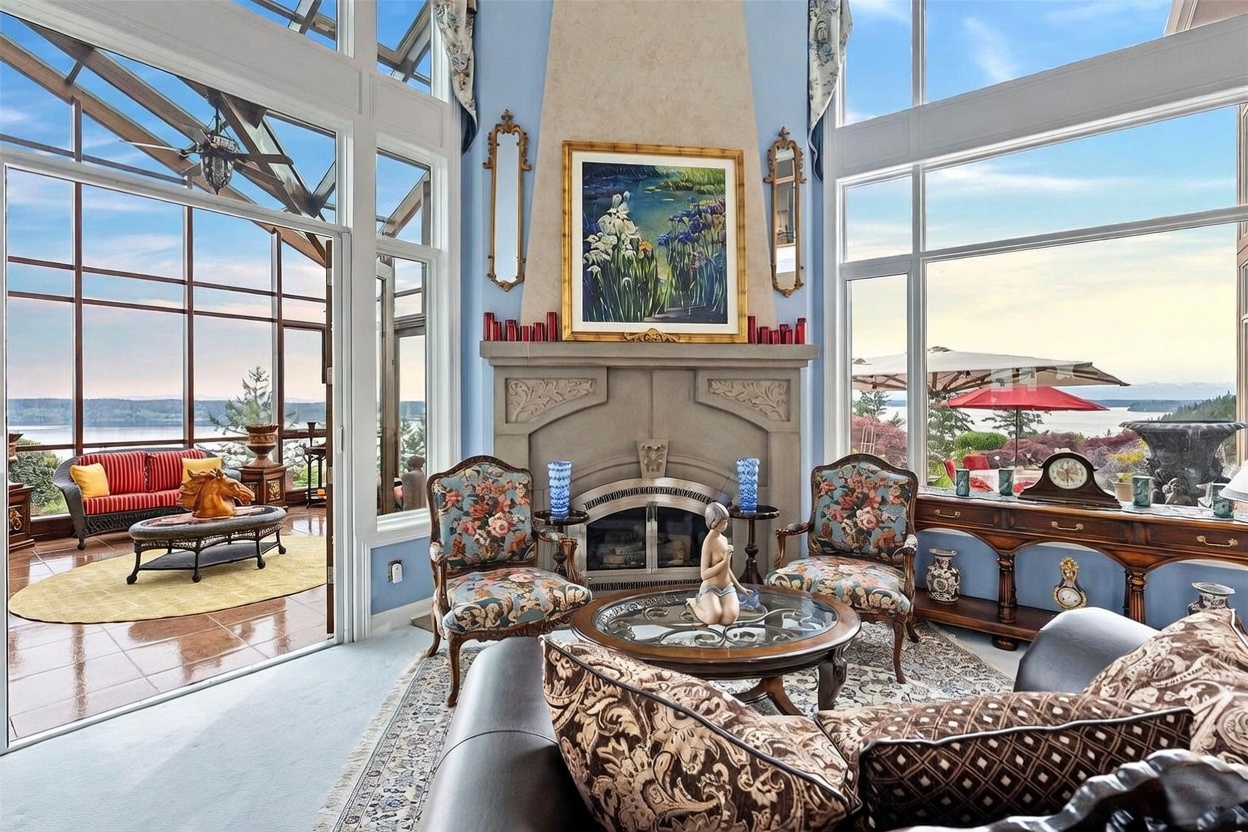 This elegant living room features a prominent stone-clad chimney breast with a central painting, flanked by tall mirrors and blue-toned walls. The space is furnished with ornate, floral-patterned armchairs and a glass-topped coffee table, all set upon a detailed area rug. Large windows provide expansive views of the water and sky, while an adjacent sunroom with a red-striped sofa is visible through a glass door.