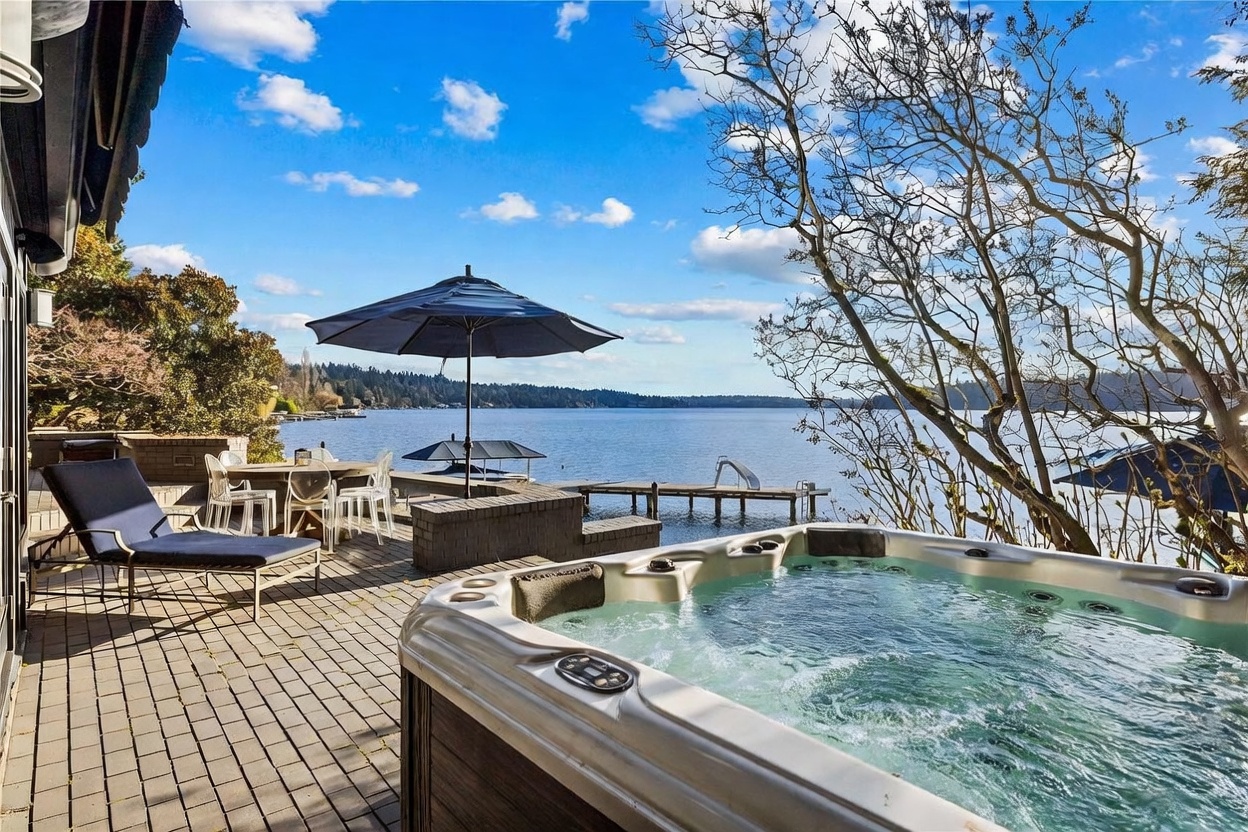 This stunning waterfront patio features a luxurious hot tub in the foreground, overlooking a serene lake with a private dock. The space is furnished with a lounge chair and an outdoor dining set under a large umbrella, creating an inviting atmosphere for relaxation and entertaining. The expansive view of the water and surrounding trees provides a peaceful, high-end outdoor living experience.