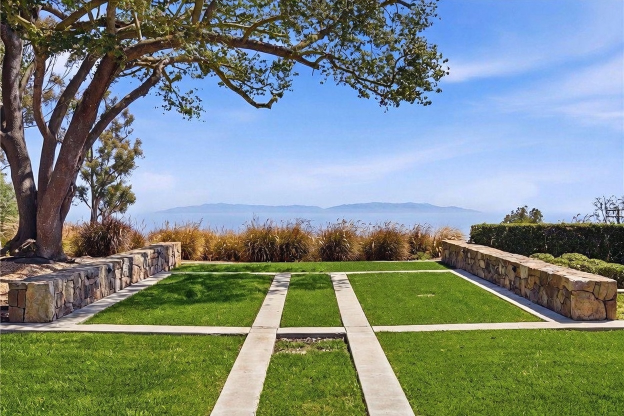 This beautifully landscaped backyard features a manicured lawn divided by concrete pathways, framed by low stone retaining walls. A large, mature tree provides shade on the left, while ornamental grasses line the perimeter, leading the eye toward a stunning, unobstructed ocean view with distant islands on the horizon. The scene is captured from a ground-level perspective, emphasizing the serene and expansive outdoor living space.