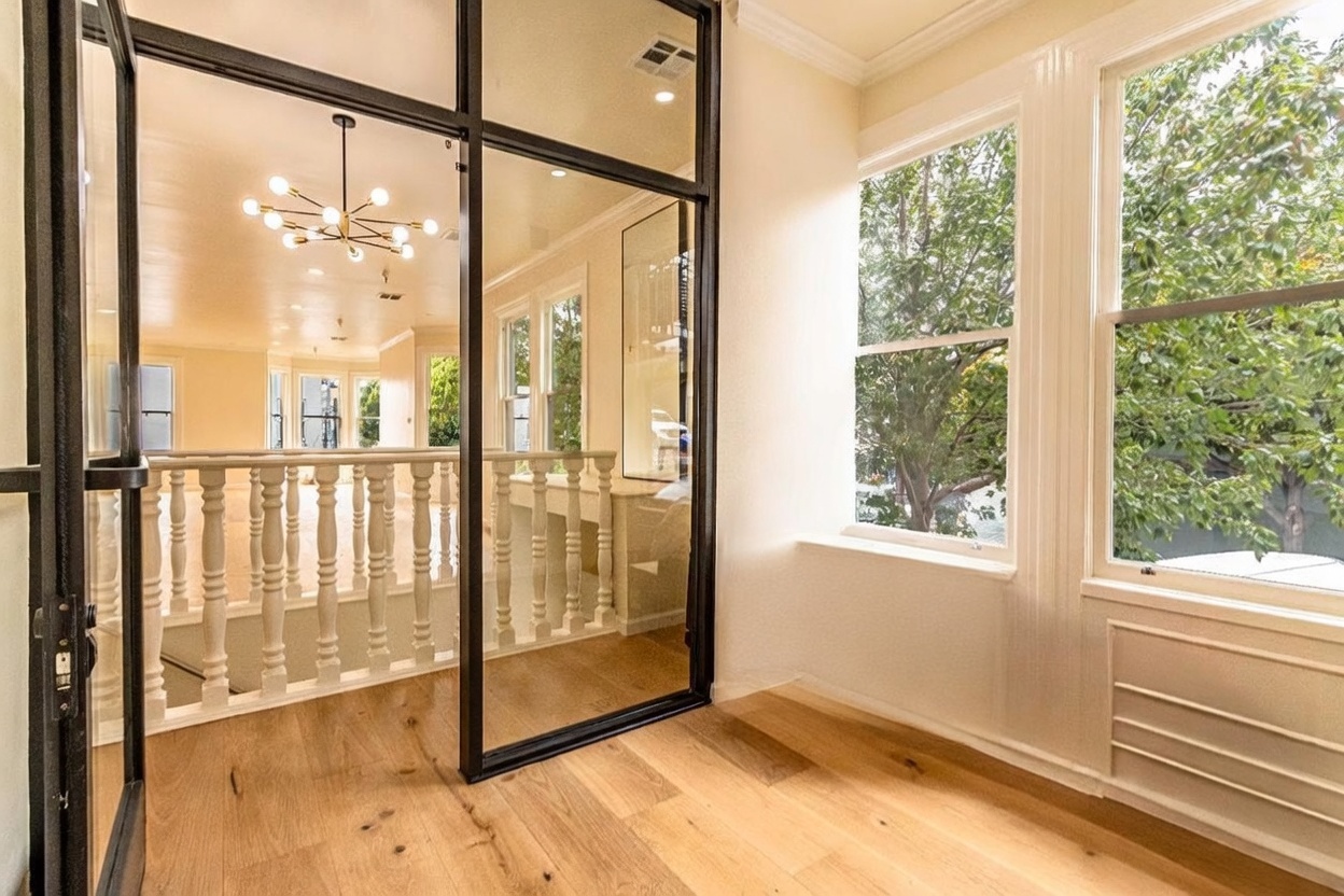 This bright sunroom features light wood flooring and large windows that offer a view of mature trees outside. A black-framed glass partition separates the space from an adjacent hallway, which is lined with a classic white balustrade and illuminated by a modern multi-arm chandelier. The room feels airy and open, creating a seamless transition between the interior living areas.