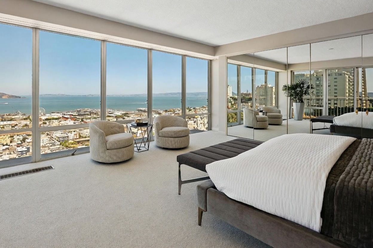 This primary bedroom features expansive floor-to-ceiling windows that provide a panoramic view of the bay and city skyline. The room is furnished with a plush bed, a dark bench at the foot, and two circular armchairs positioned near the glass. A large mirrored wall reflects the interior space, enhancing the sense of openness and light throughout the room.