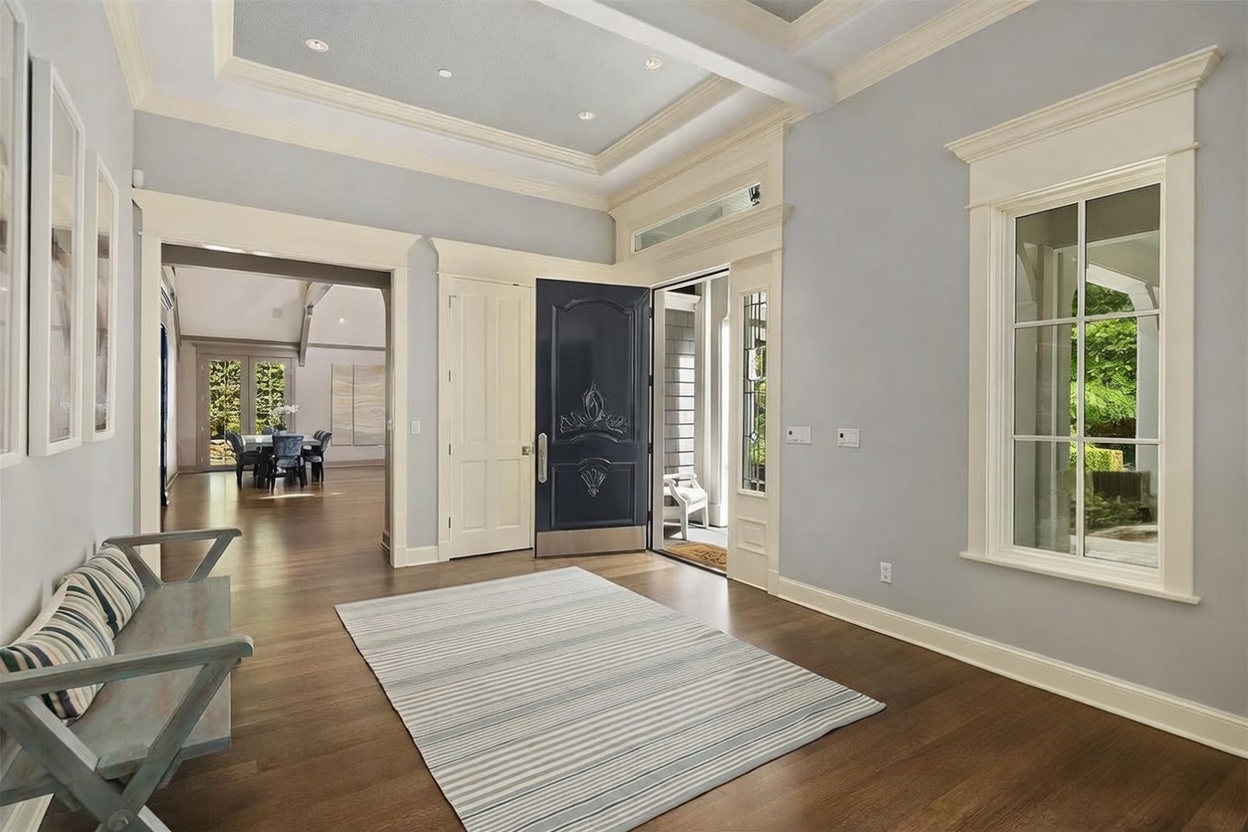 This elegant foyer features a dark blue front door with ornate detailing, set against light gray walls and crisp white trim. A striped area rug sits on the dark wood flooring, complemented by a light-colored armchair and a wooden bench with striped cushions. The space is brightened by a coffered ceiling with recessed lighting and a large window that allows natural light to pour into the room.