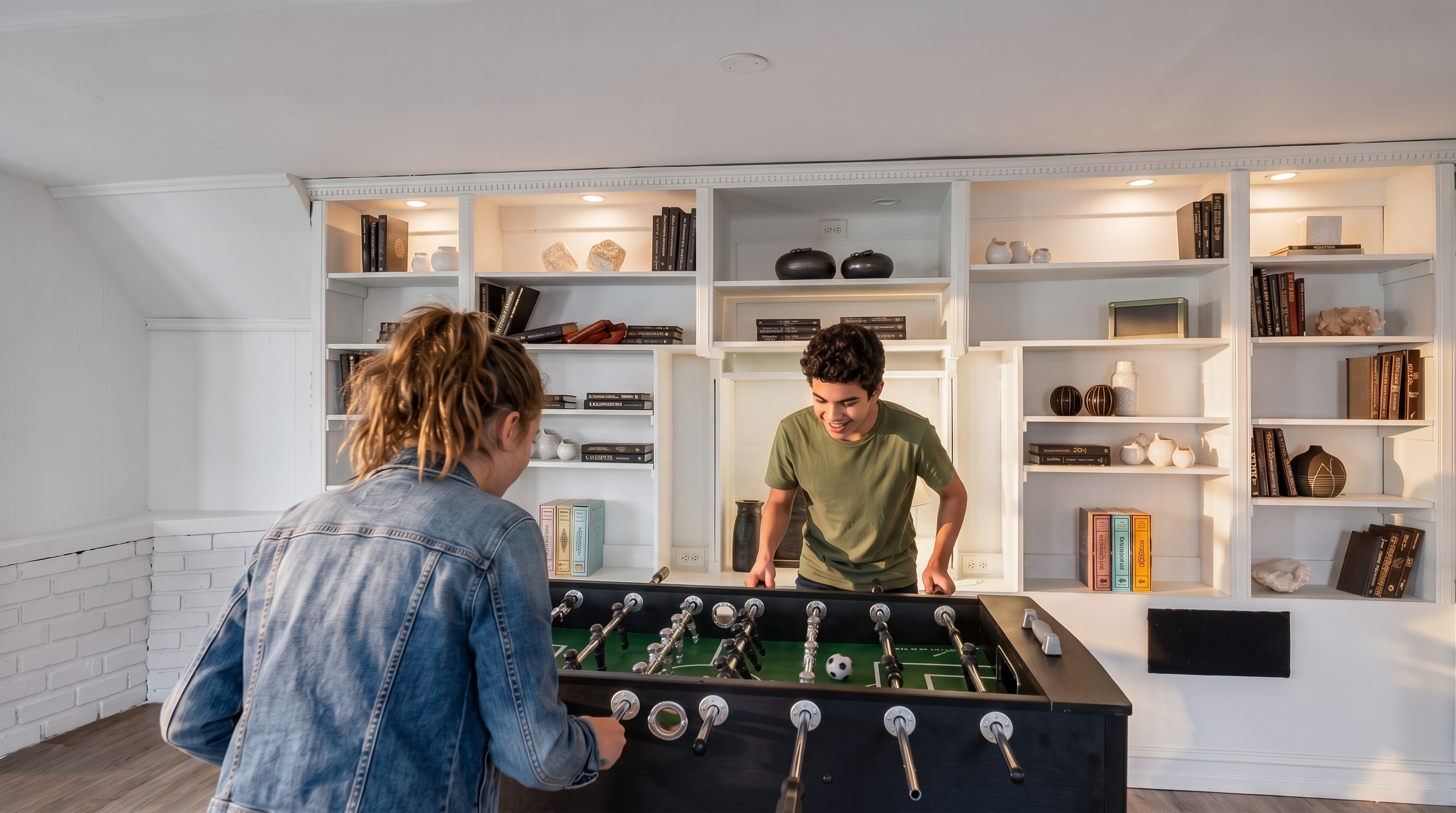 This recreational space features a prominent black foosball table positioned in the foreground, set against a large, custom-built white shelving unit that spans the wall. The shelving is styled with a mix of books, decorative vases, and board games, creating a curated and organized look. The room has a casual, inviting atmosphere, with light-toned wood flooring and a white-painted brick wall section on the left, suggesting a finished basement or game room setting.