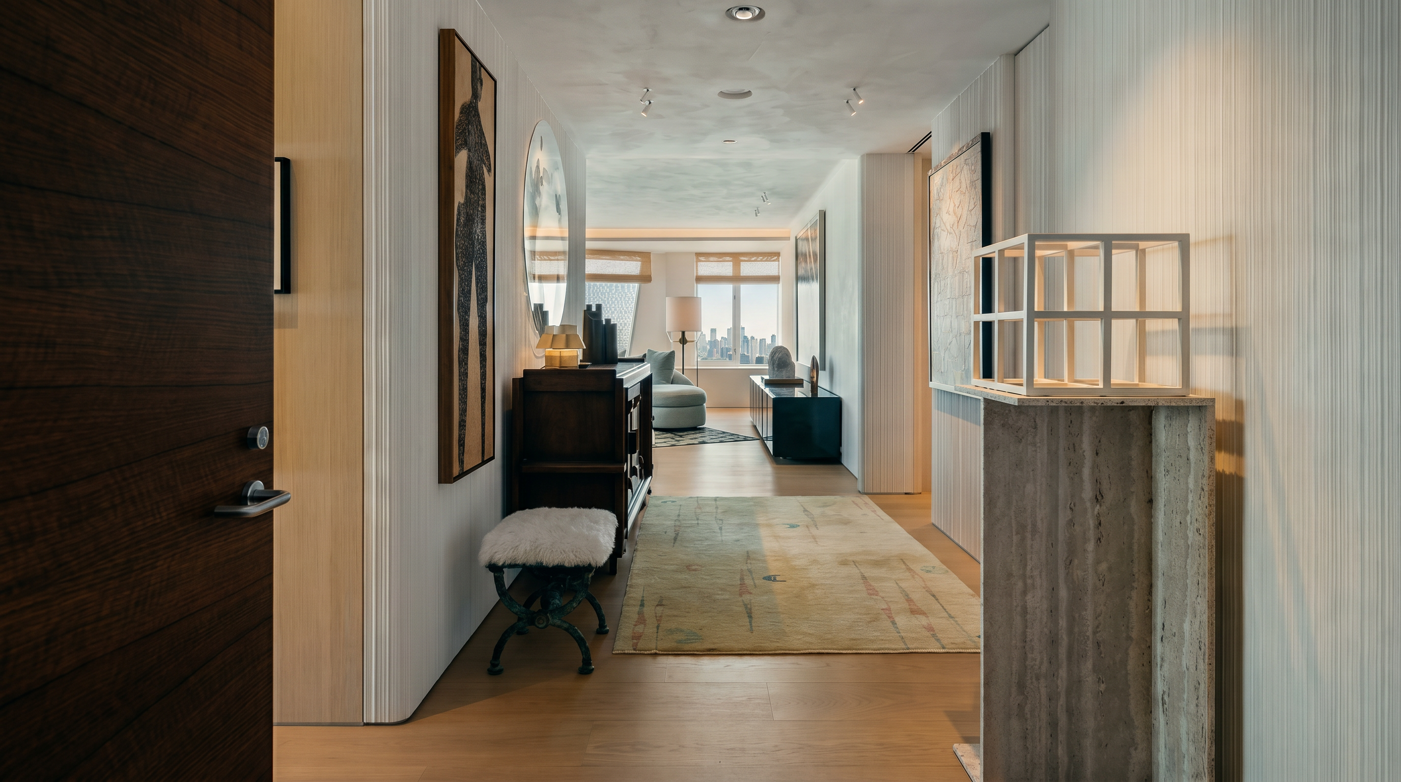 This elegant entryway and hallway feature light wood flooring and textured wall coverings, creating a sophisticated and airy atmosphere. A sculptural stone pedestal with a geometric wire frame sits in the foreground, while a dark wood console table and a plush, fur-topped stool add texture and warmth. The perspective looks down the gallery-style hall toward a bright living area, emphasizing the open and refined layout of the home.