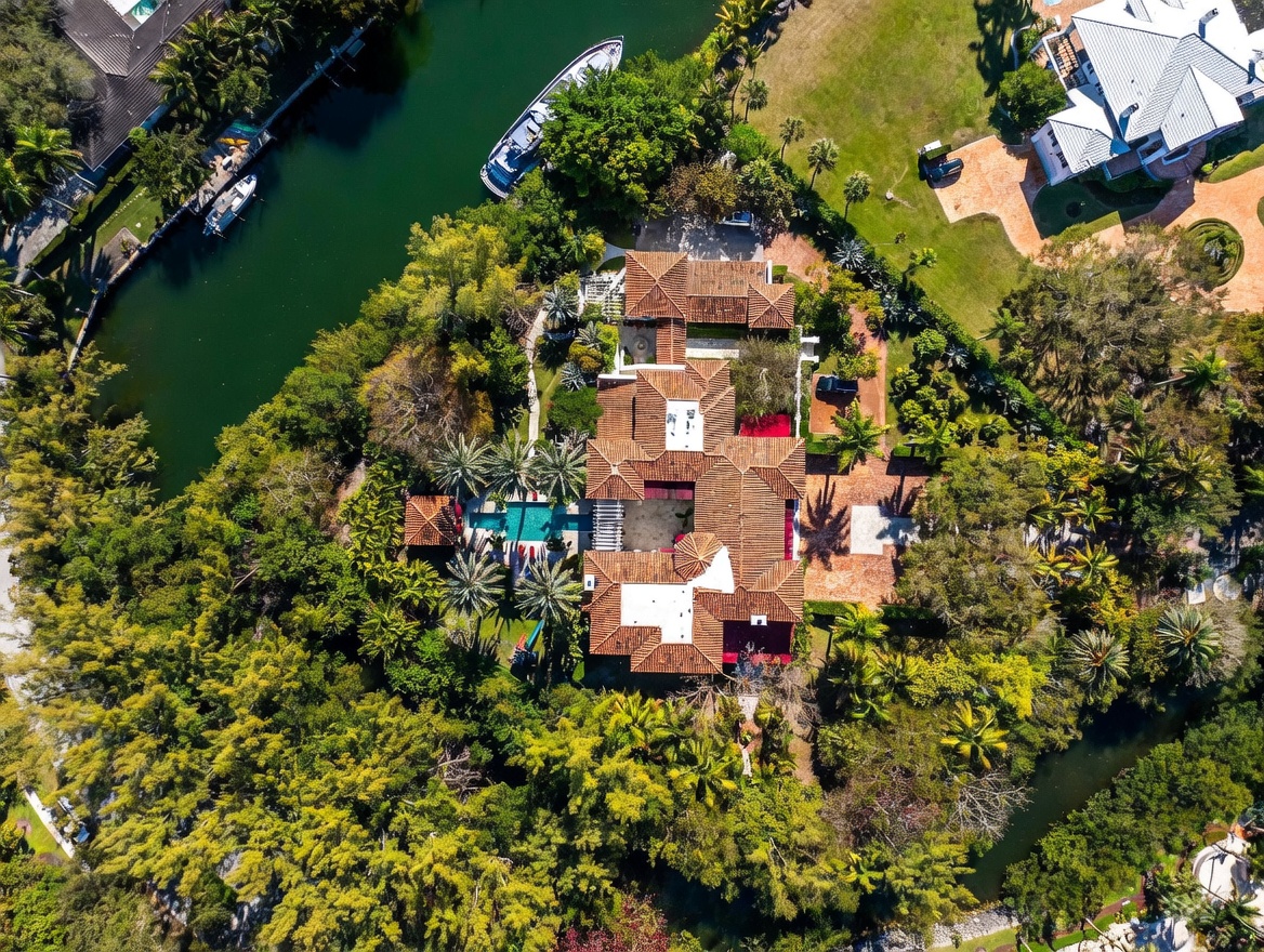 This high-angle aerial view captures a sprawling Mediterranean-style estate nestled within a lush, tree-filled landscape. The property features a complex roofline with terracotta tiles, a private swimming pool, and extensive brick-paved courtyards, all situated along a winding canal with docked boats. The dense surrounding vegetation and waterfront location provide a secluded and luxurious atmosphere.