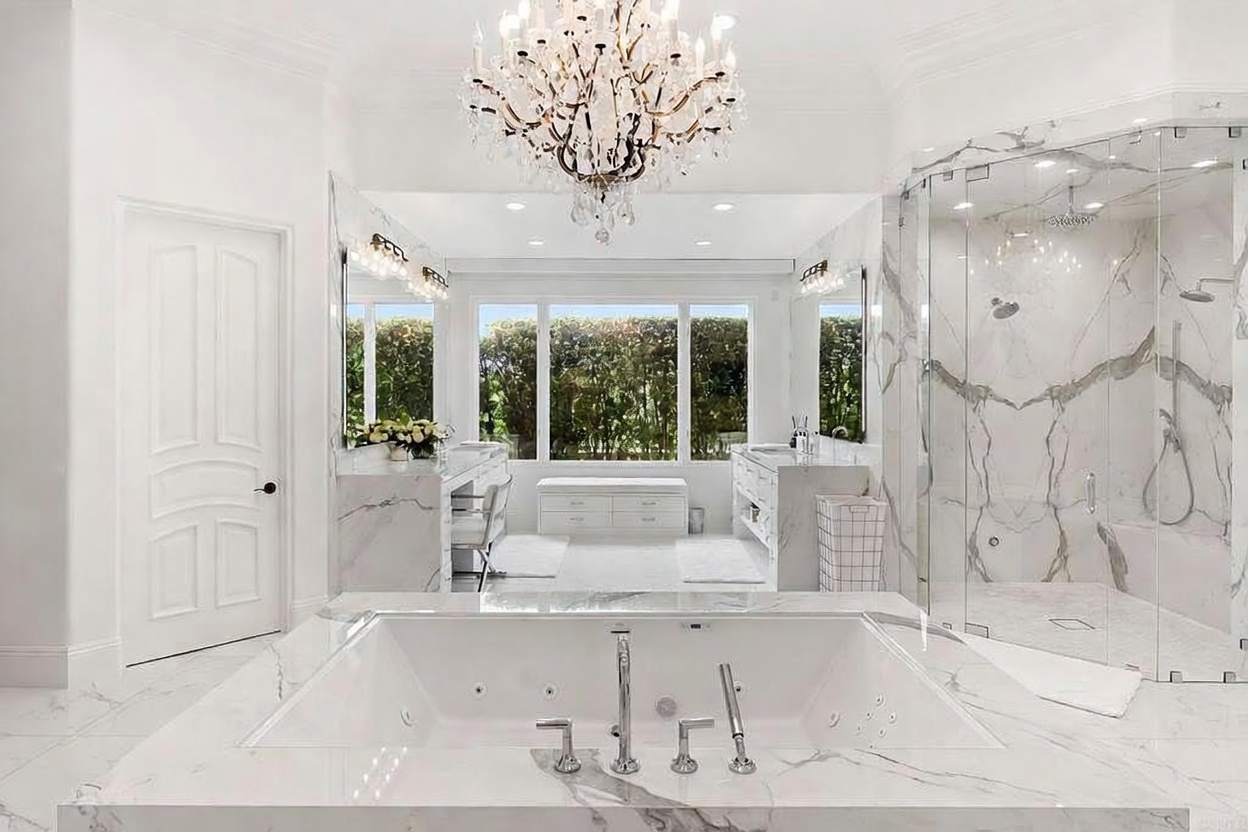 This is a luxurious primary bathroom featuring a large soaking tub in the foreground, surrounded by marble flooring and walls. A stunning chandelier hangs above, and the room includes dual vanities with mirrors, a separate glass-enclosed shower, and large windows providing natural light. The overall impression is one of elegance and spa-like tranquility.