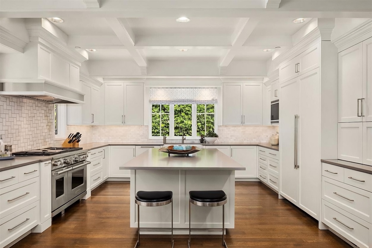 This bright, spacious kitchen features crisp white cabinetry, a large central island with seating, and high-end stainless steel appliances. The space is anchored by rich wood flooring and a classic herringbone-patterned backsplash, while a coffered ceiling adds architectural interest above. A large window over the sink provides natural light, creating an inviting and clean aesthetic perfect for modern living.
