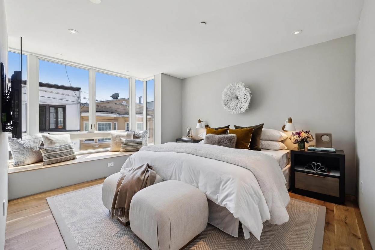 This bright and airy primary bedroom features a large bed centered against a light gray wall, accented by a decorative wall hanging and dual wall-mounted sconces. A spacious window seat with plush cushions provides a cozy nook overlooking the neighborhood, while light wood flooring and a textured area rug add warmth to the space. The room is completed by a dark wood nightstand and two upholstered ottomans at the foot of the bed, creating a sophisticated and inviting atmosphere.