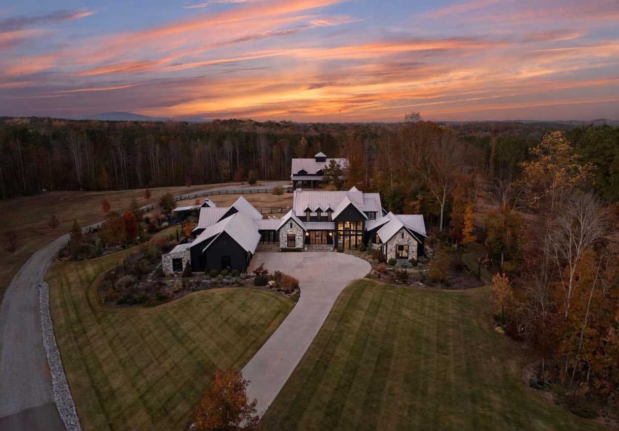 This stunning aerial perspective captures a sprawling, modern farmhouse-style estate set against a vibrant sunset sky. The home features a complex roofline with metal roofing, dark exterior siding, and stone accents, complemented by a circular driveway and manicured lawn. The property is surrounded by dense, mature trees, creating a private and serene atmosphere that highlights the home's architectural grandeur.
