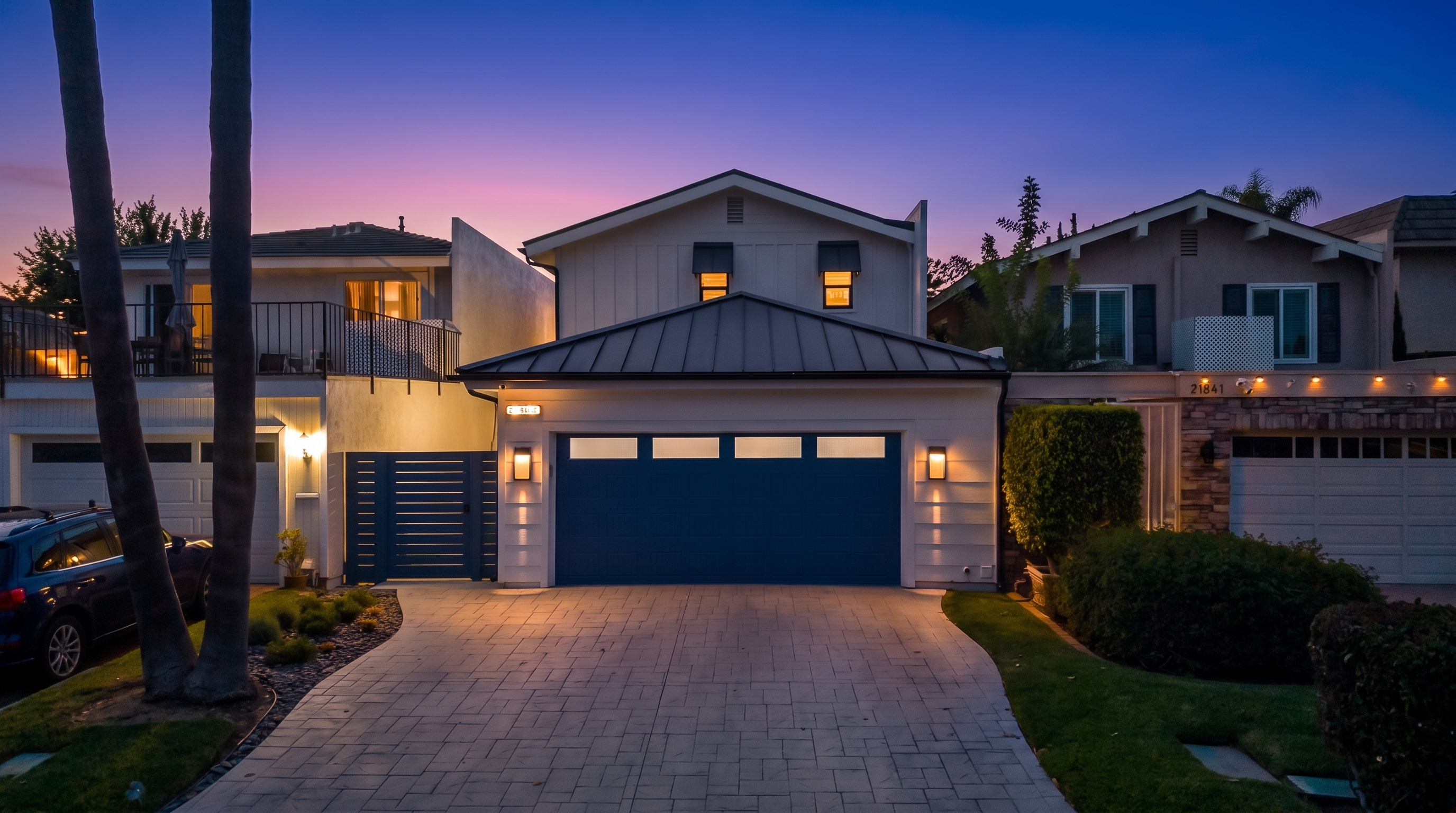 This modern suburban home features a clean white facade with vertical board-and-batten siding and a prominent dark blue garage door. The property includes a neatly paved driveway, subtle architectural lighting, and two small upper-level windows with black awnings, creating a polished and inviting curb appeal at dusk.